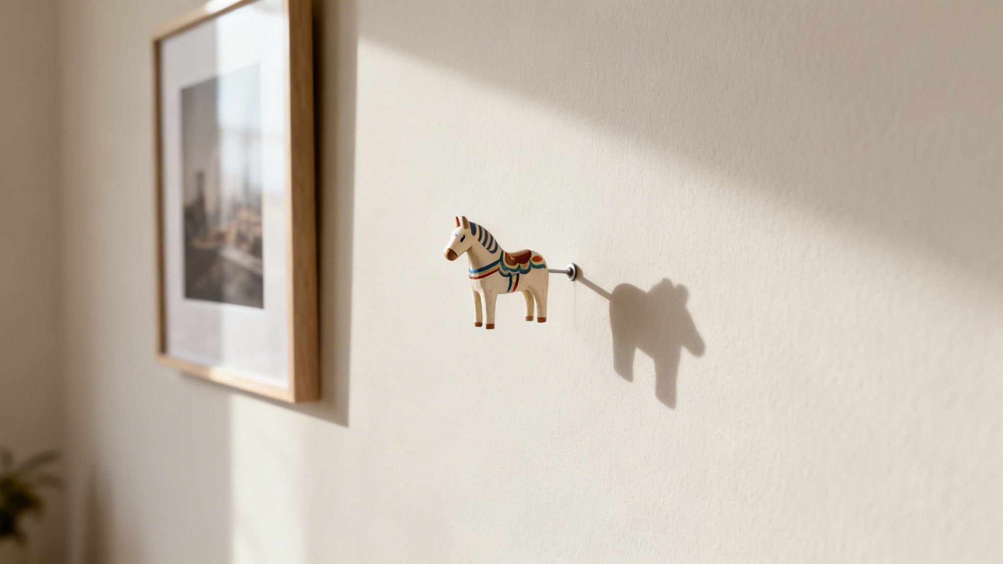A small, colorful wooden horse pin casts a distinct shadow on a bright wall, next to a blurred framed picture.