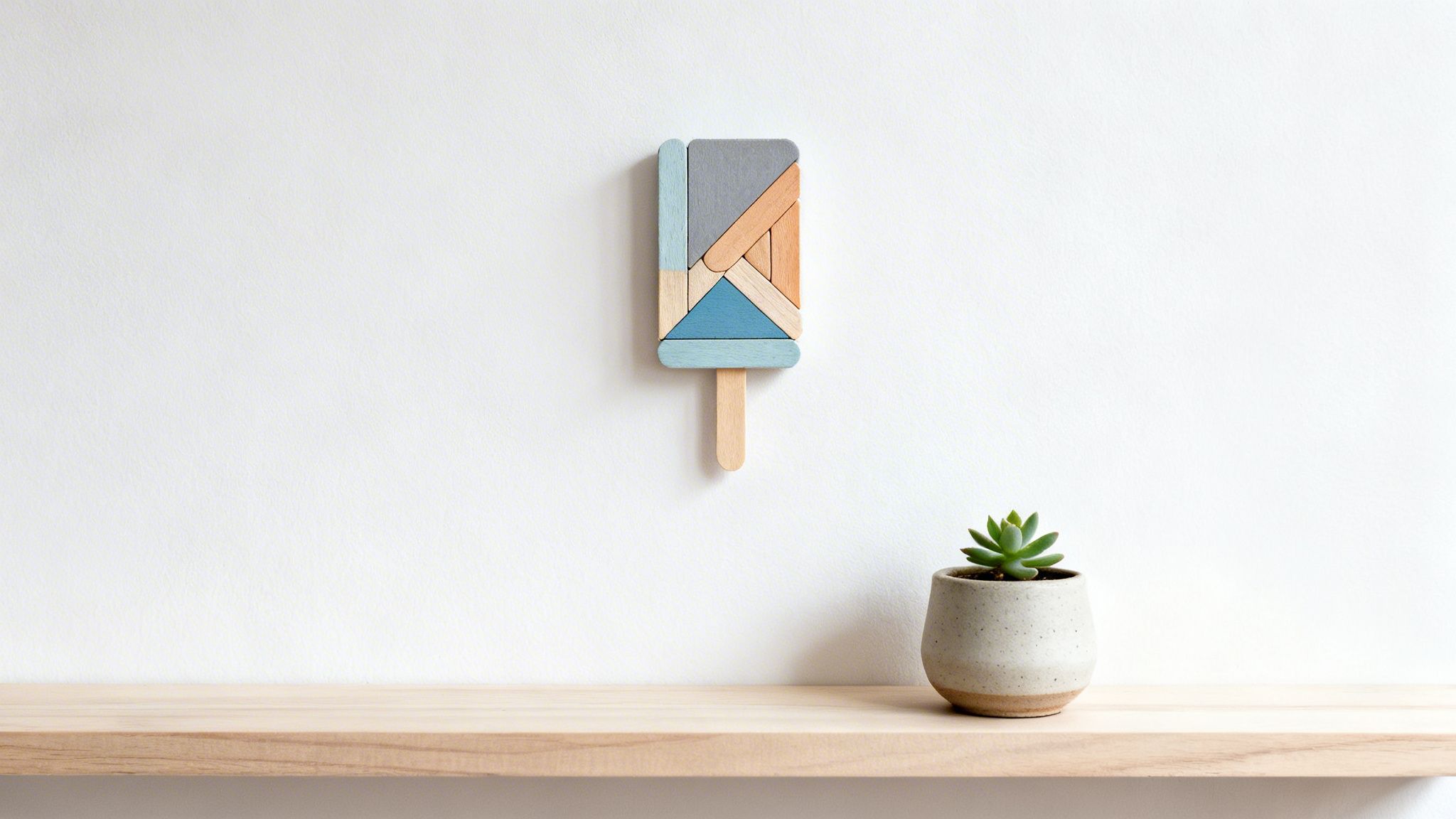 A minimalist white wall with a wooden ice pop art piece and a succulent plant on a shelf.