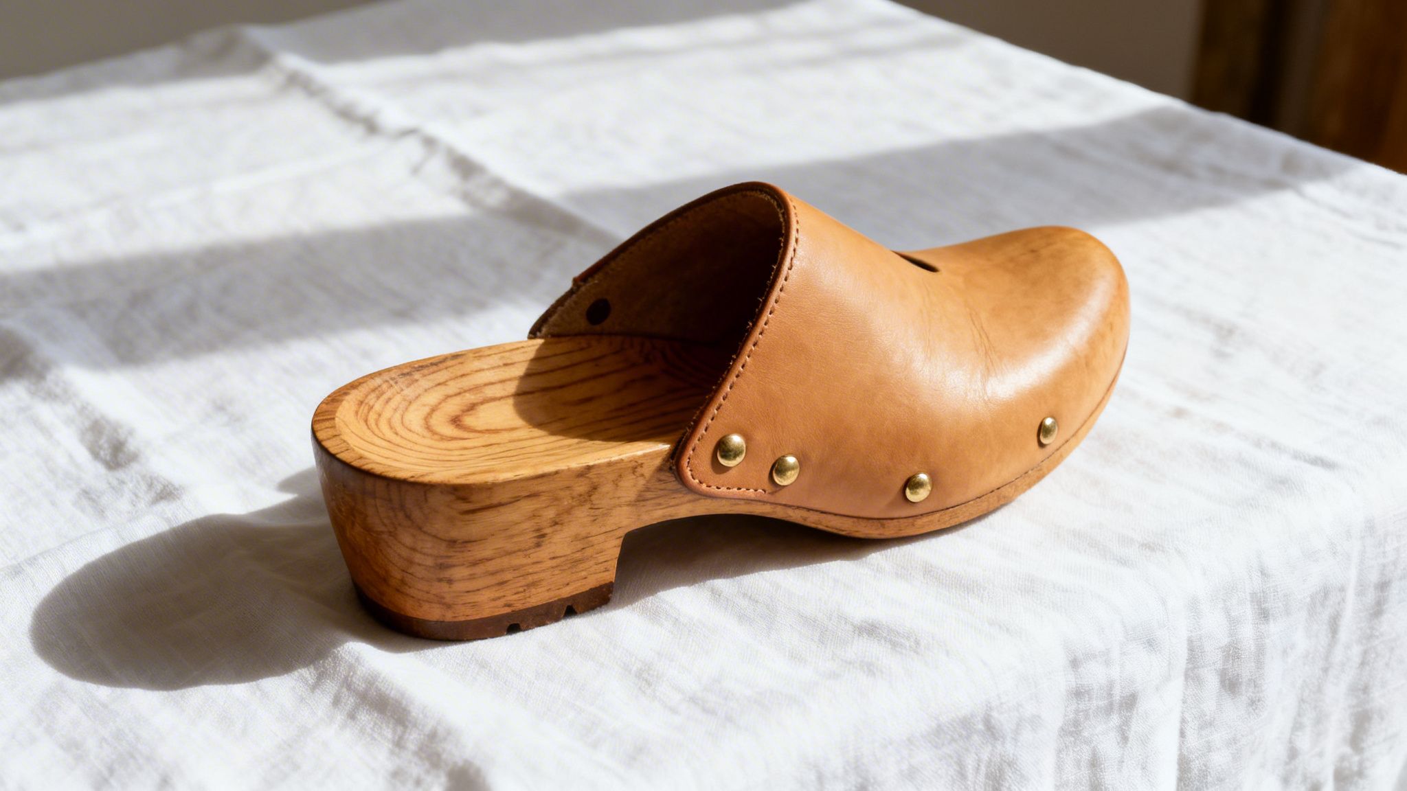 A single light brown leather clog with a wooden sole and brass studs, sitting on white fabric.