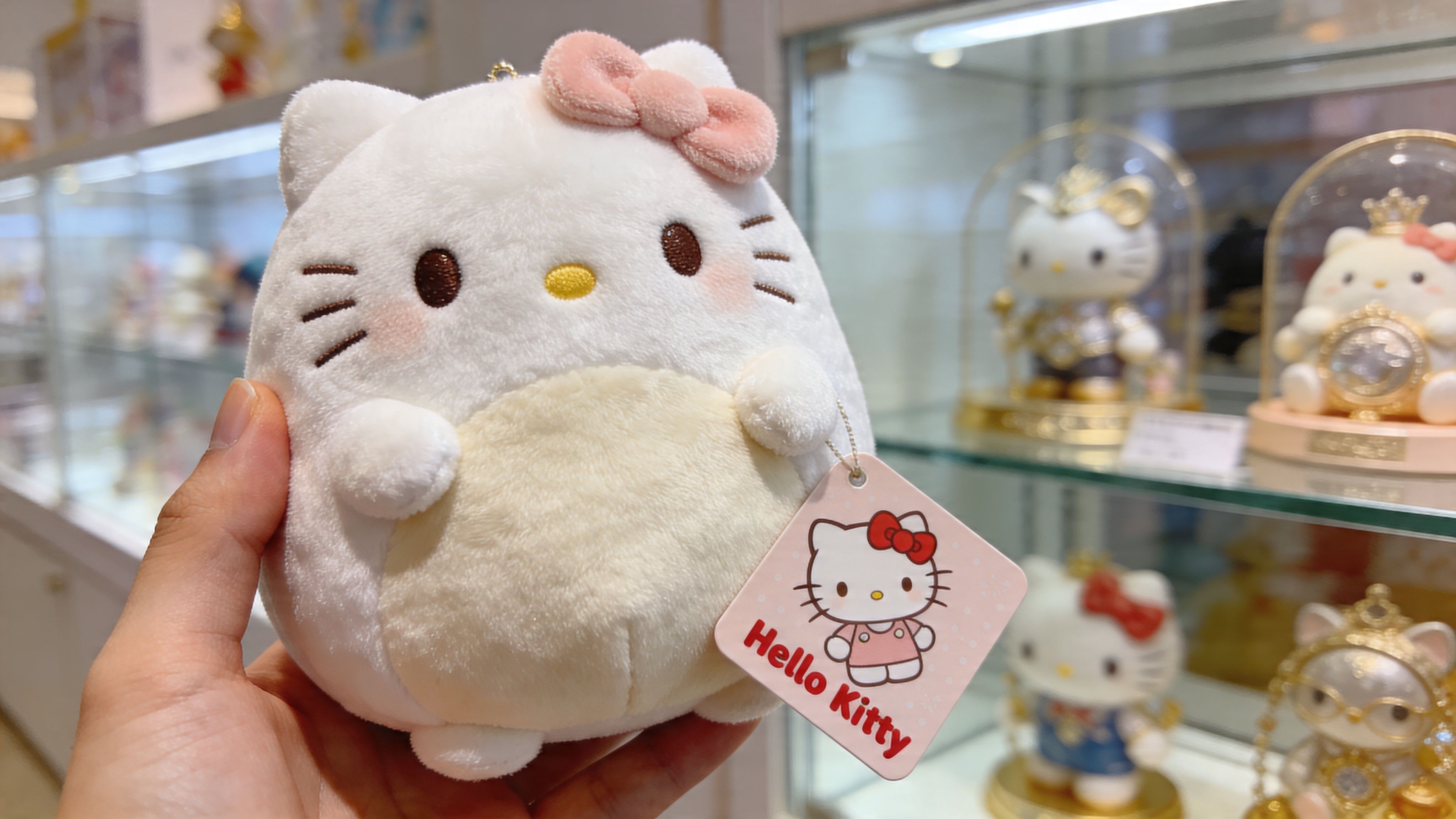 A hand holds a plush Hello Kitty Squishmallow with a pink bow and a hanging brand tag.