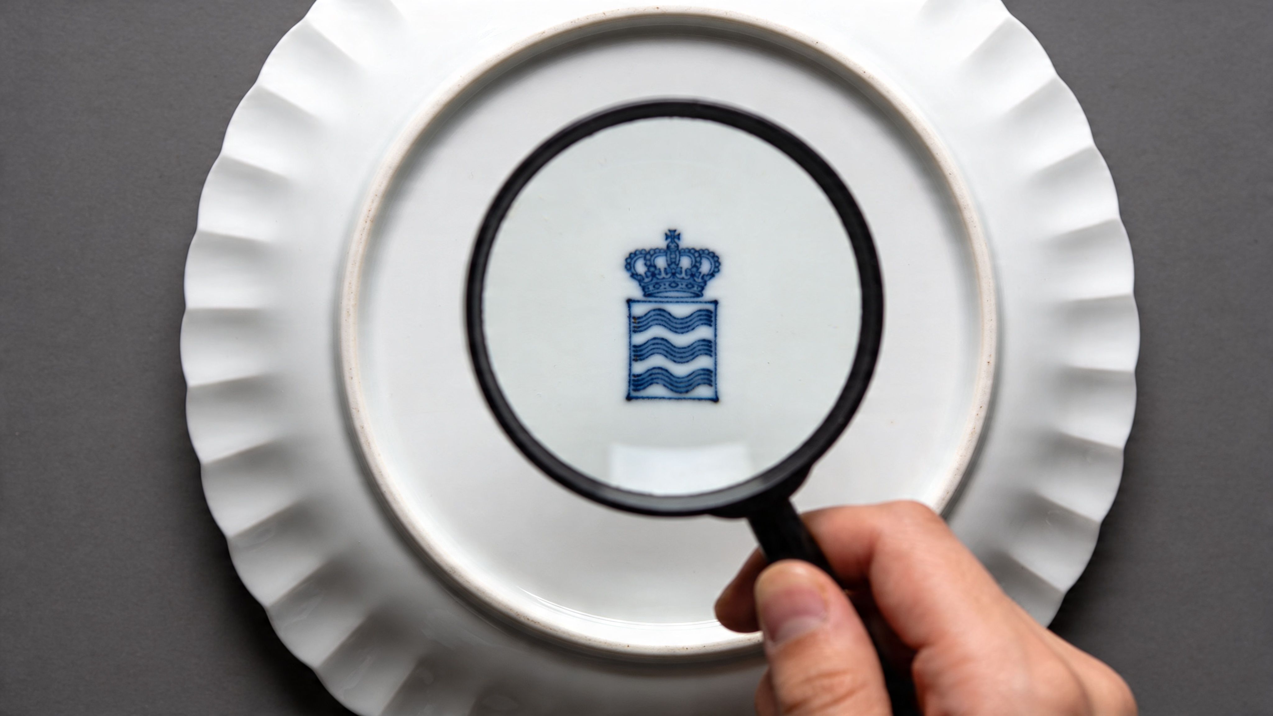 A hand holding a magnifying glass over a blue stamped porcelain mark on a white plate.