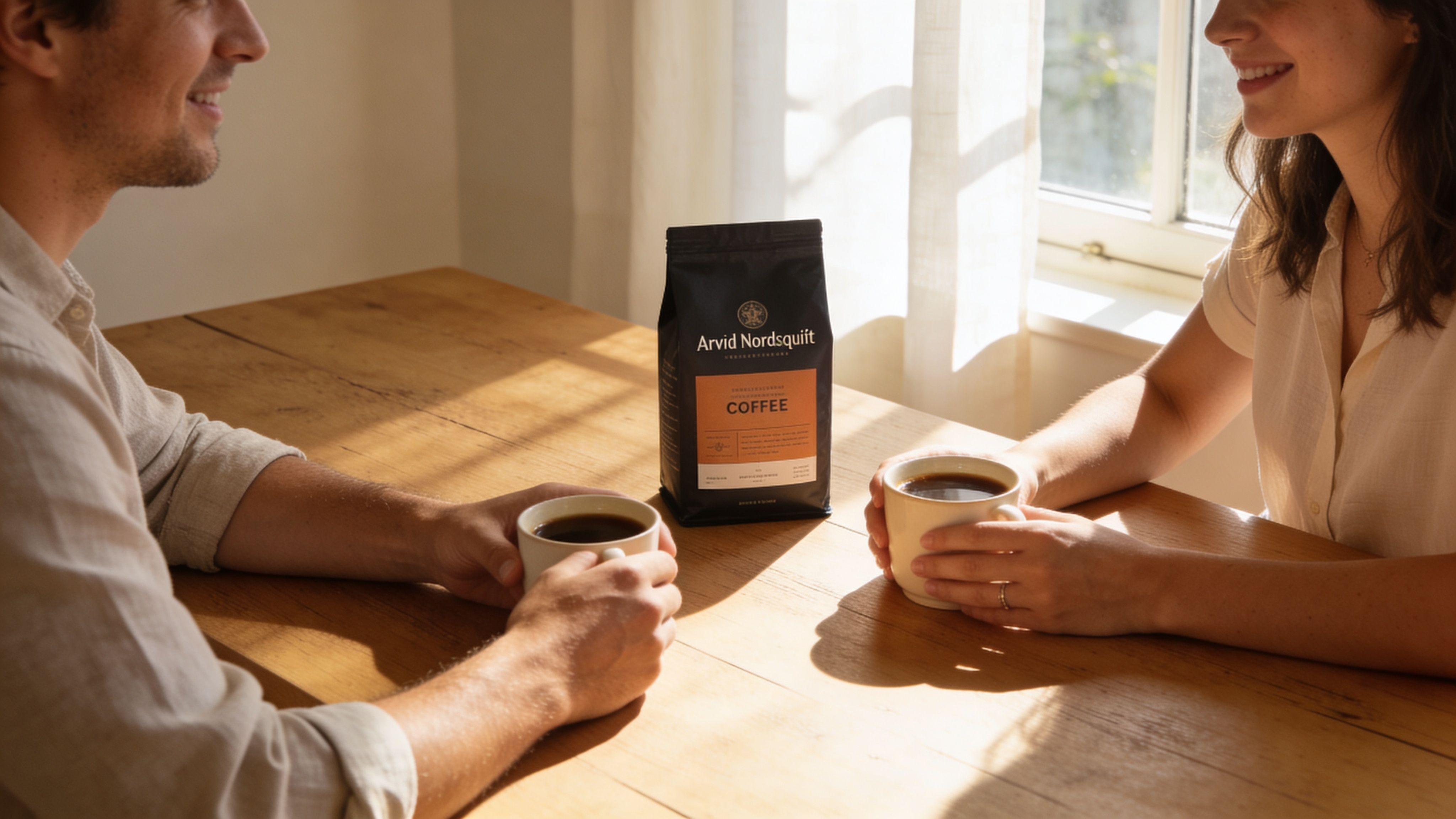 A man and a woman sitting at a wooden table smiling while holding mugs of coffee.