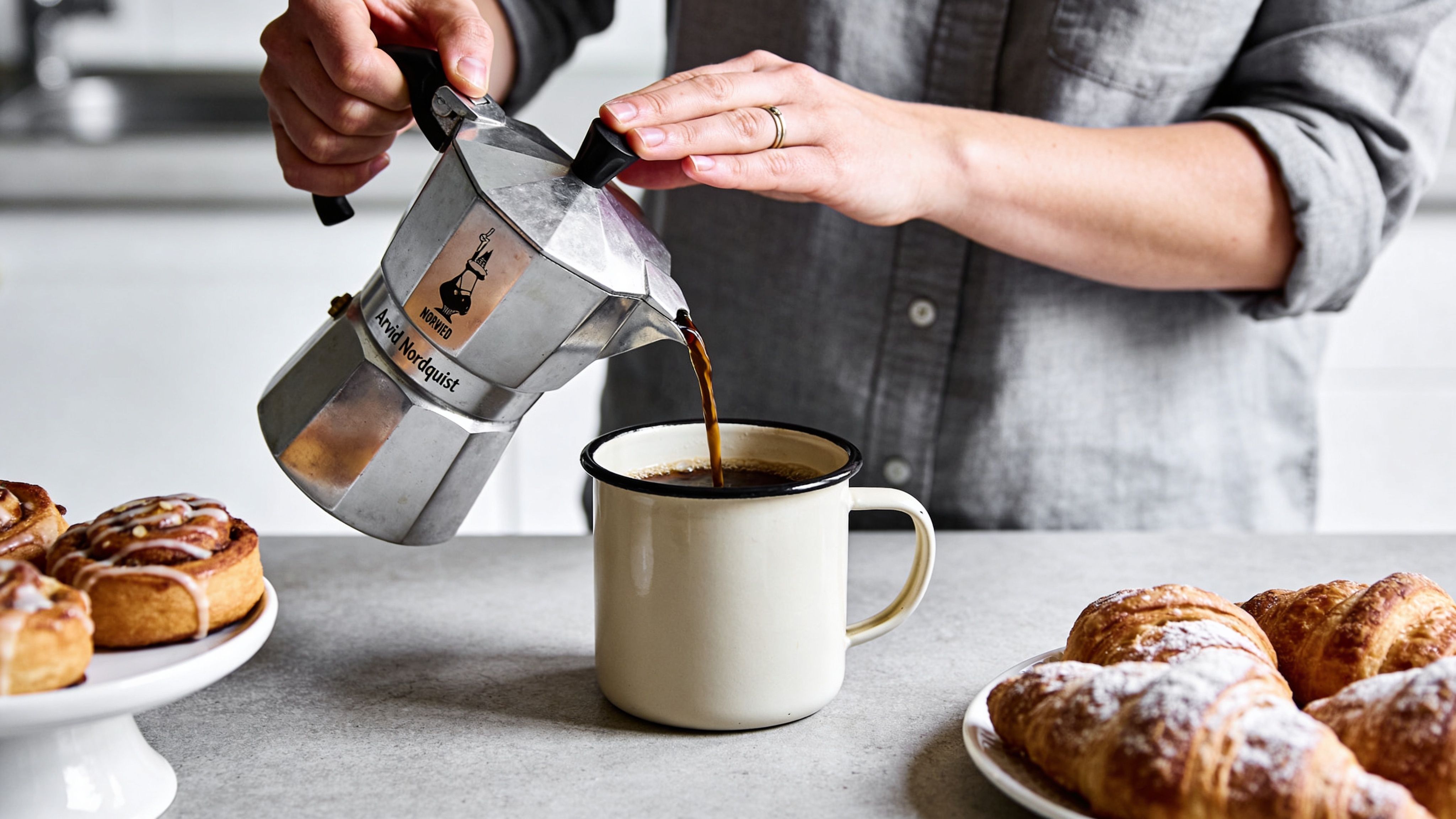 A person pouring dark coffee from an Arvid Nordquist moka pot into a white enamel mug.