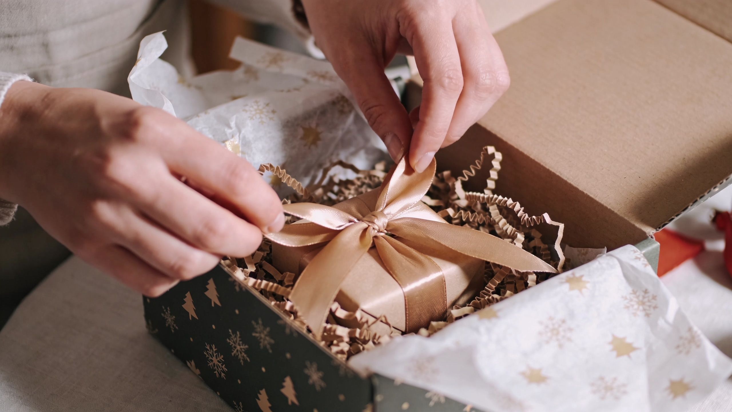A person carefully tying a gold ribbon on a small gift box inside a decorated holiday gift box.