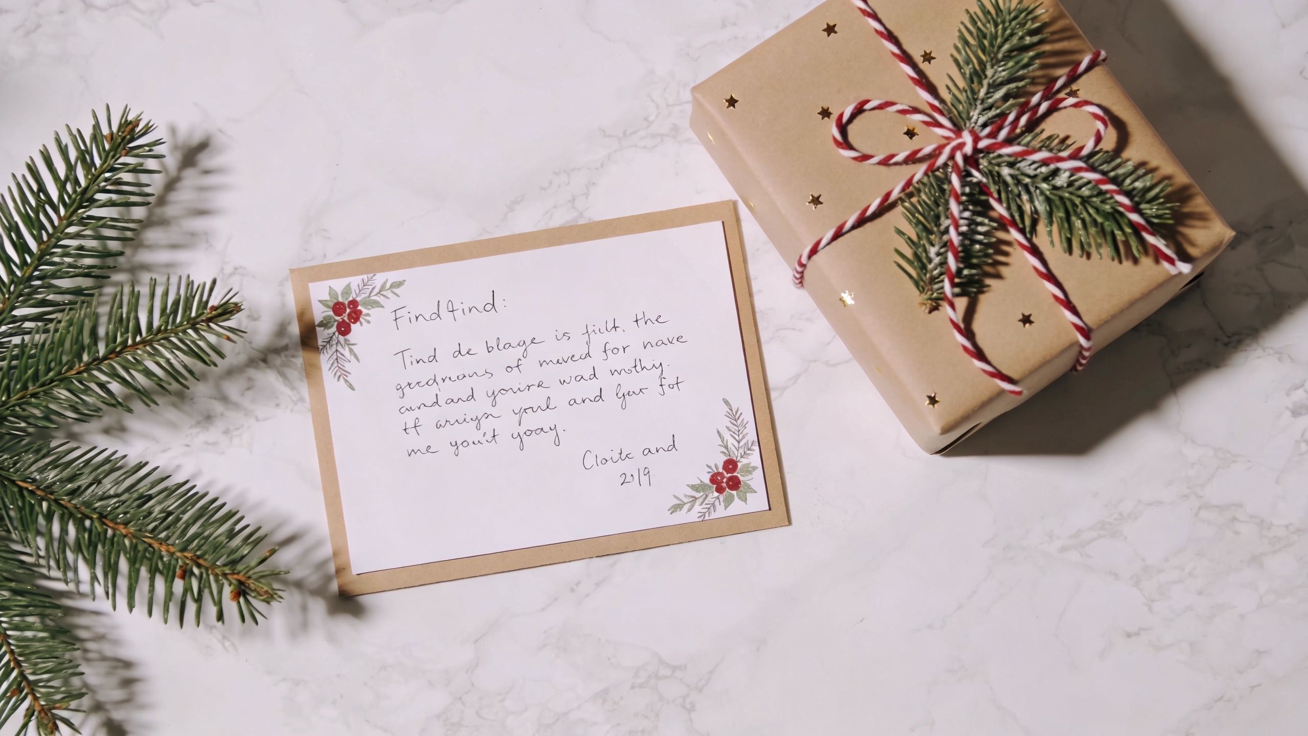 A brown gift box decorated with greenery and a handwritten Christmas card on a white marble surface.