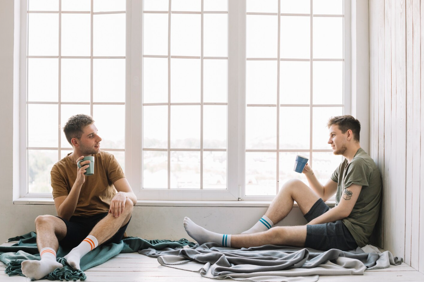 Two men sit on a windowsill in a recovery housing living space, holding mugs and talking in a relaxed, sunlit environment that reflects peer support and community