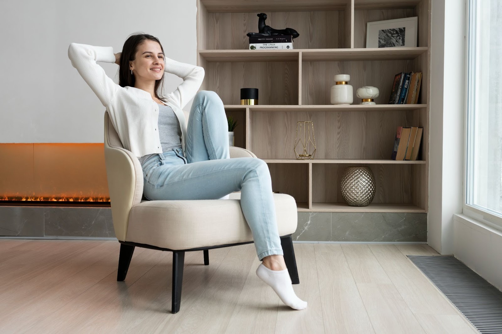 A woman in casual clothing relaxes on a beige armchair in a calm recovery housing living space, smiling with her hands behind her head near a modern fireplace