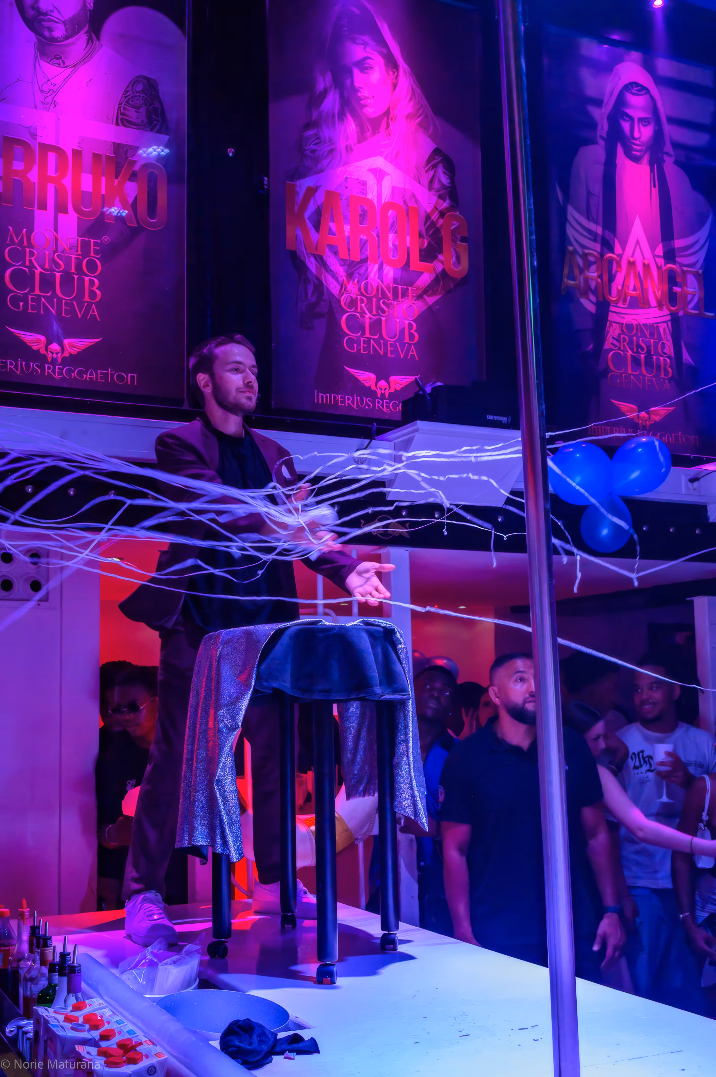Noé fait voler des banderoles de confettis tout autour de lui pendant un spectacle au Montécristaux Club, à Genève. Devant lui se trouve une table ayant servi à faire voler quelqu’un, ainsi qu’une barre de pole dance. En bas de l’image, on aperçoit des bouteilles et des boissons qui seront servies aux clients visibles sur la droite de l’image.