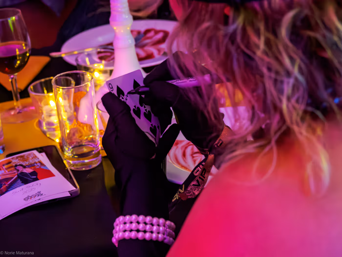 Une personne portant des gants noirs et un bracelet de perles signe une carte à jouer 9 de pique dans un cadre festif aux éclairages colorés avec des verres et une coupe de vin sur la table.