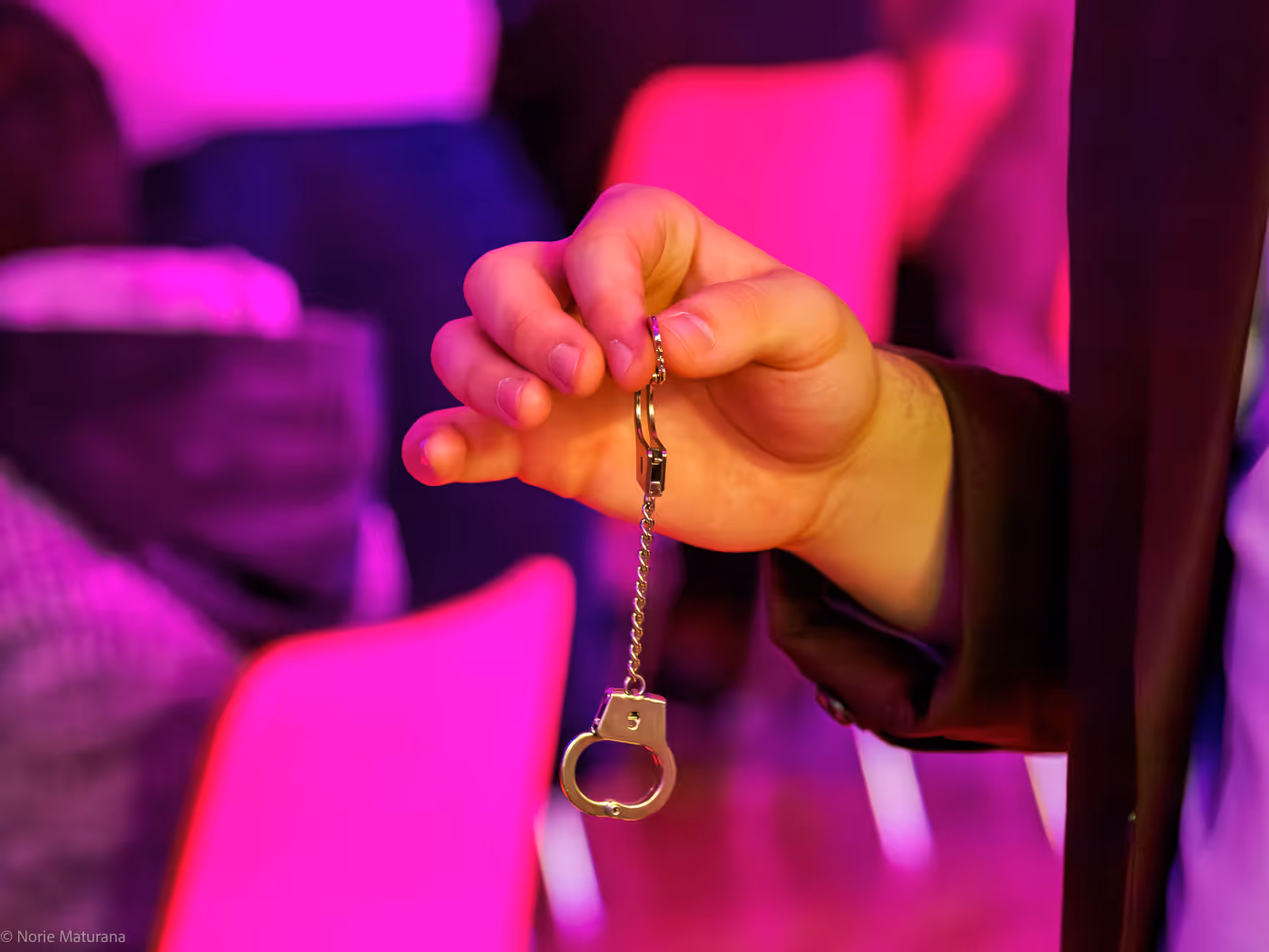 Main tenant une menotte miniature en métal suspendue à une chaîne dans un environnement éclairé de rose.