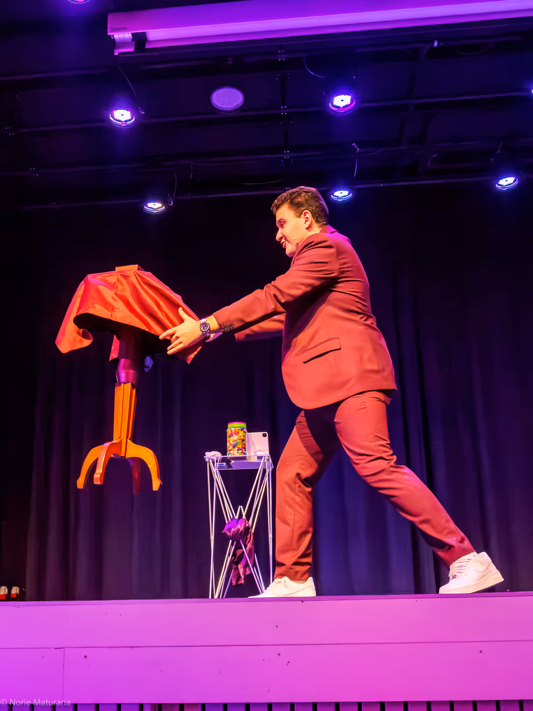 Magicien en costume bordeaux effectuant un tour avec une petite table recouverte d'un tissu rouge sur scène éclairée en violet.