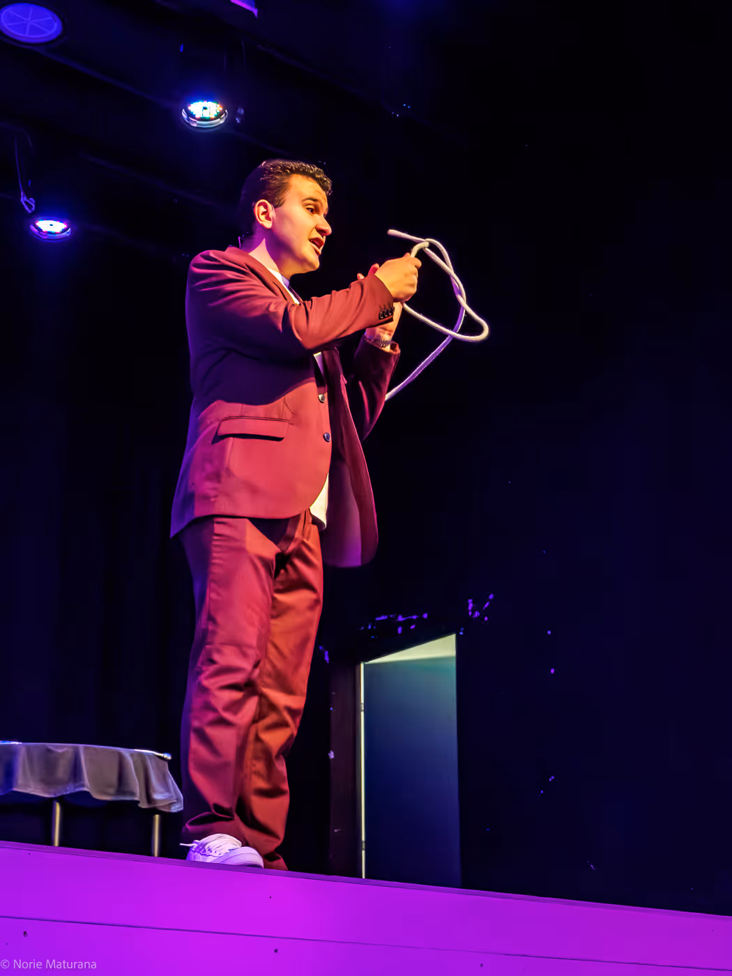 Un magicien sur scène en costume bordeaux manipule une corde dans la lumière violette.