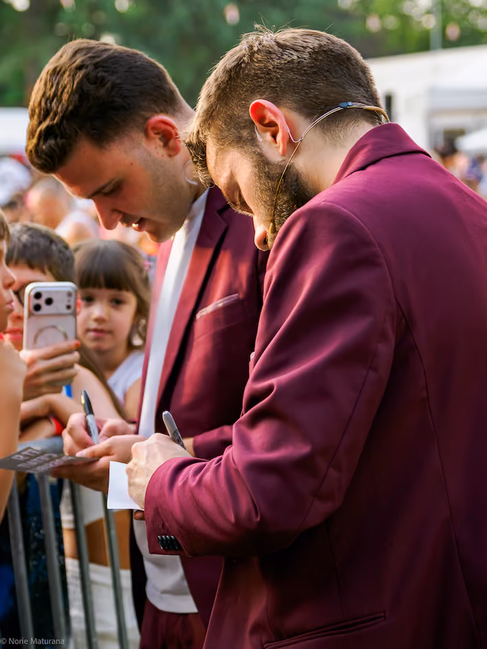 Deux magiciens en vestes bordeaux signent des autographes pour des enfants derrière une barrière.