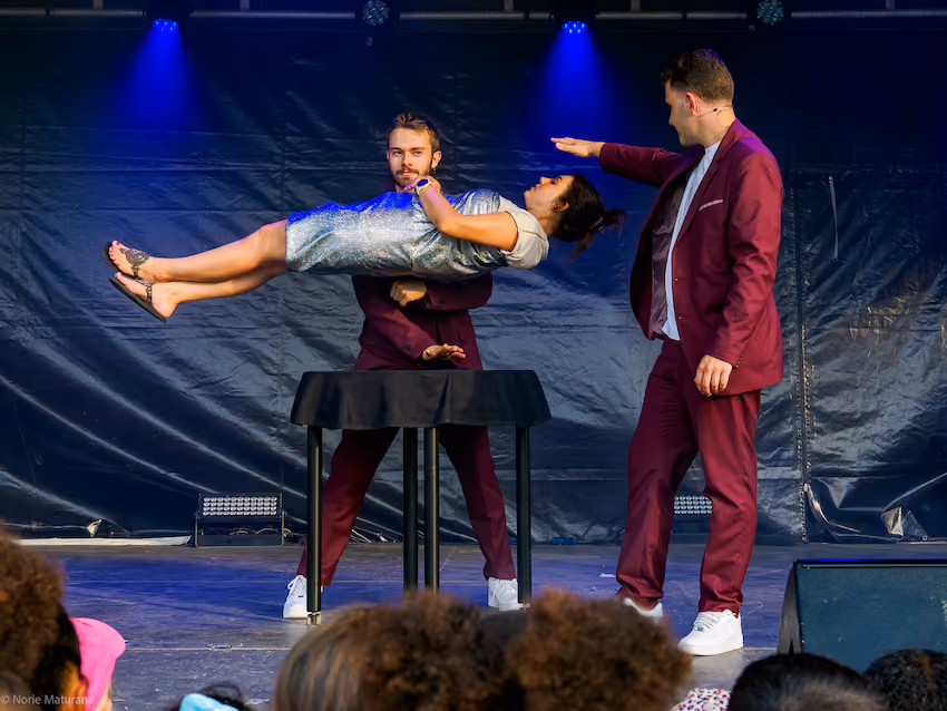 un magicien en costume bordeaux réalise un tour de magie avec une femme flottant horizontalement au-dessus d'une table.