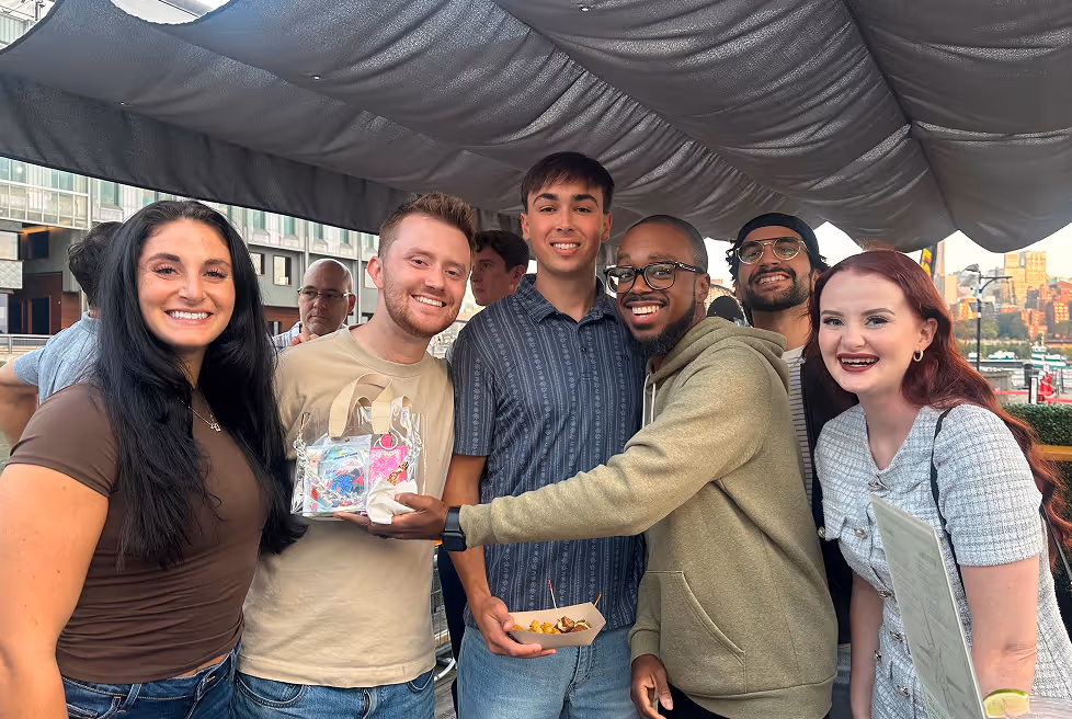 Group of six young adults smiling and posing together under a canopy in an outdoor urban setting, with one person holding a clear gift bag and another holding a food tray.