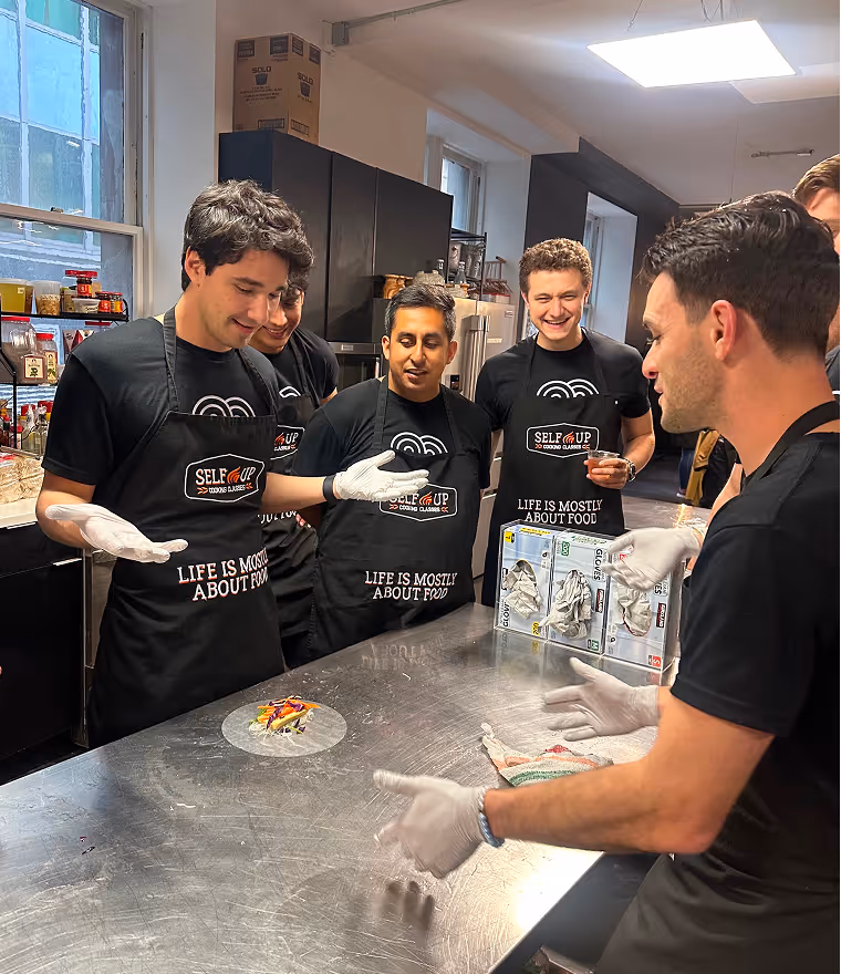 Group of men wearing 'Self Up Cooking Classes' aprons and gloves gathered around a kitchen counter with a small plated dish.