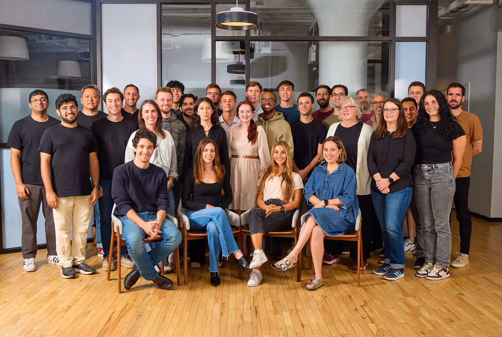 Large group of diverse men and women posing together indoors on wooden floor, some seated on chairs and others standing behind.