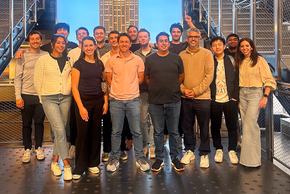 Group of sixteen diverse adults posing together on a modern industrial-style balcony with a tall building in the background.