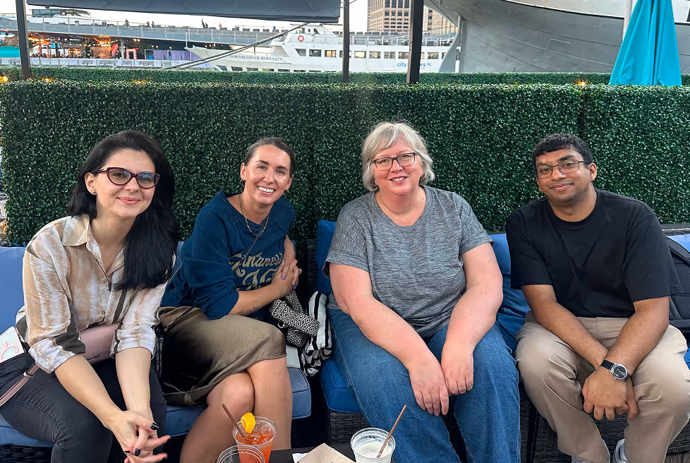 Four people sitting on blue outdoor seats in front of green hedge, smiling at the camera with drinks on a table in front.