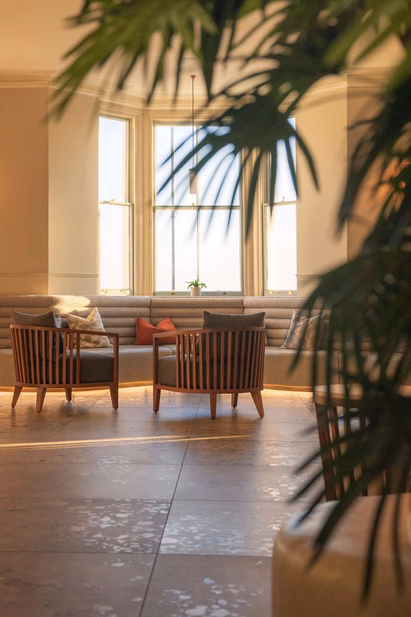 Sunlit lounge area at The Esplanade hotel, Llandudno, with tables and chairs beside tall windows, viewed through green foliage