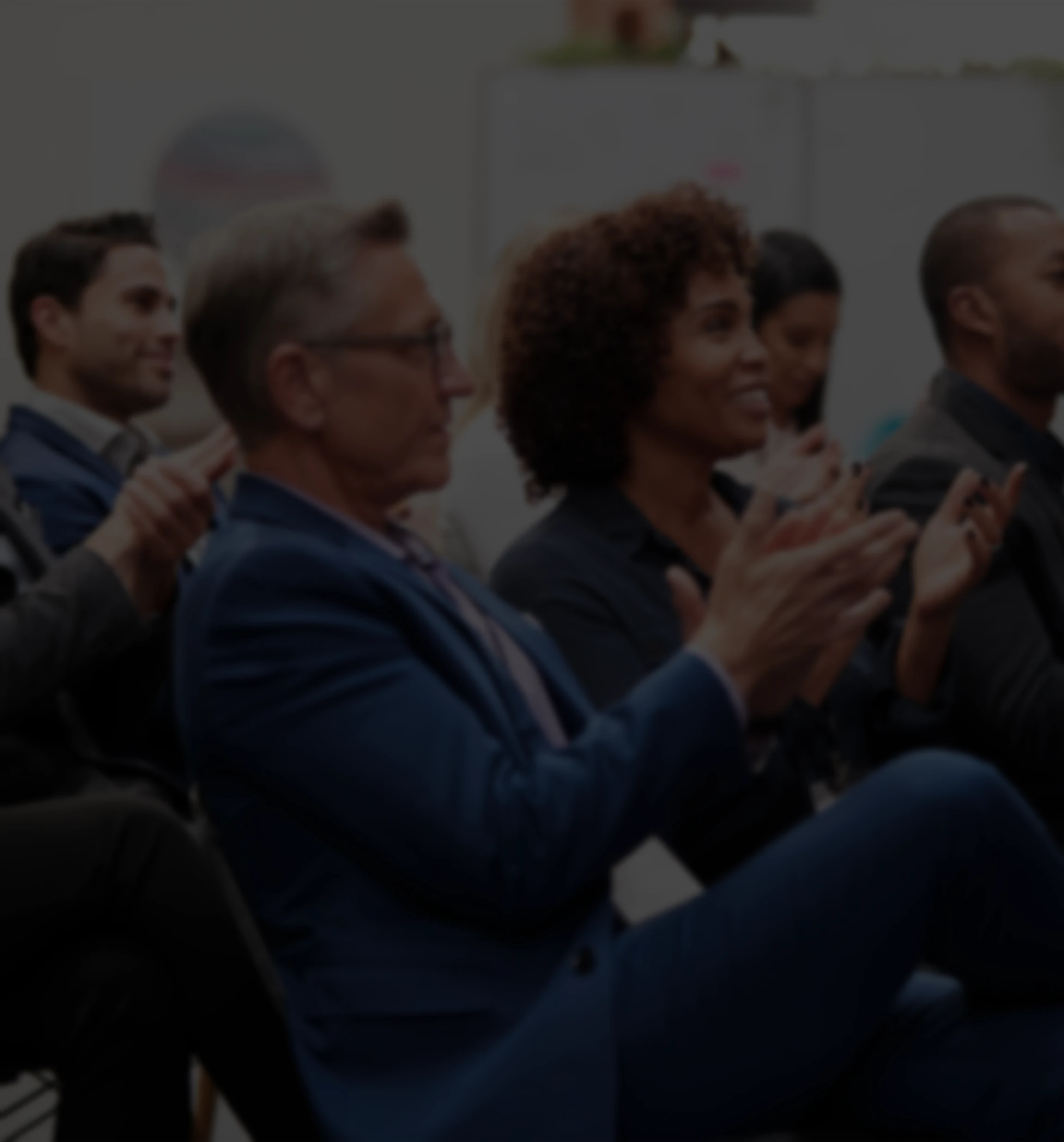 A diverse audience in a dark conference room clapping during a presentation.