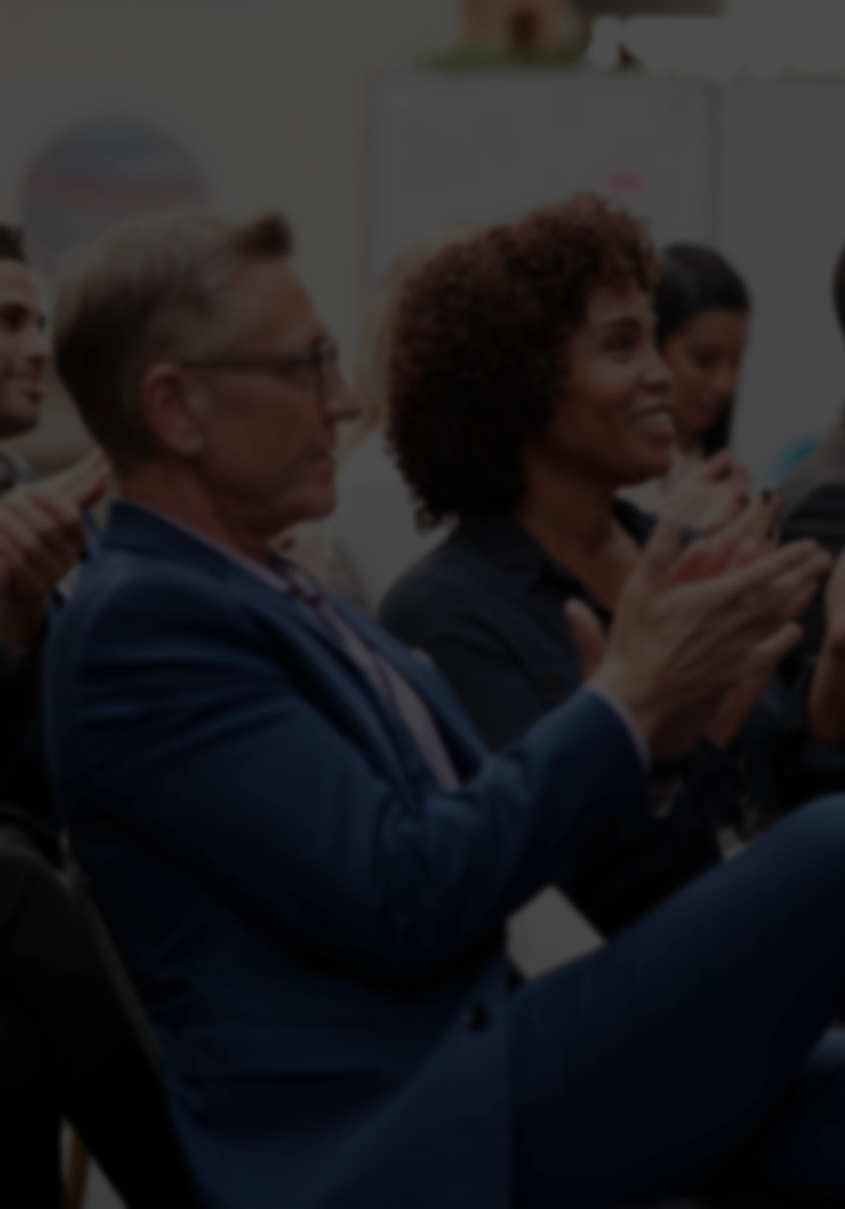 A diverse audience in a dark conference room clapping during a presentation.