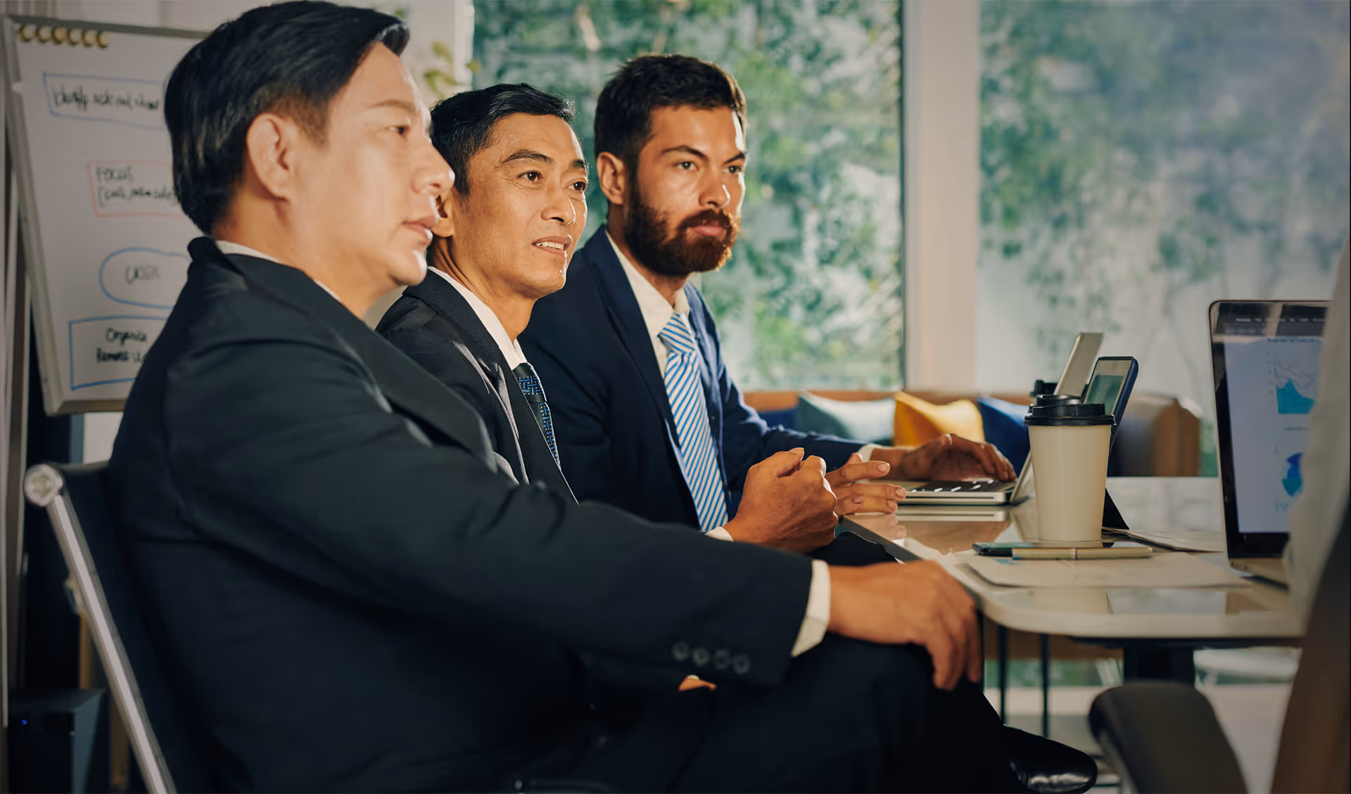 Three businessmen in suits sitting at a conference table, engaged in a meeting with laptops, documents, and a coffee cup visible.