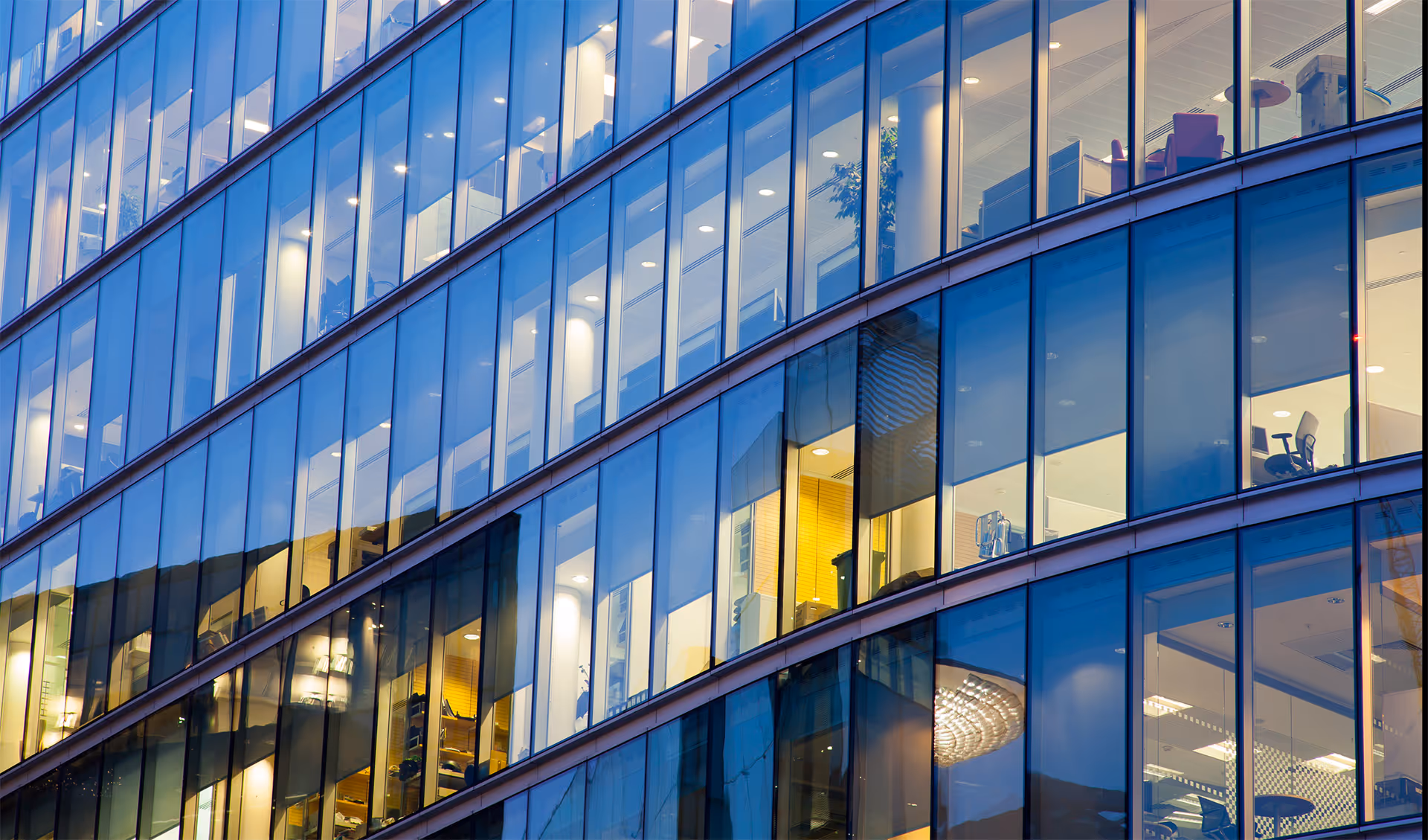 Curved glass facade of an office building with illuminated interior and visible desks, chairs, and plants.