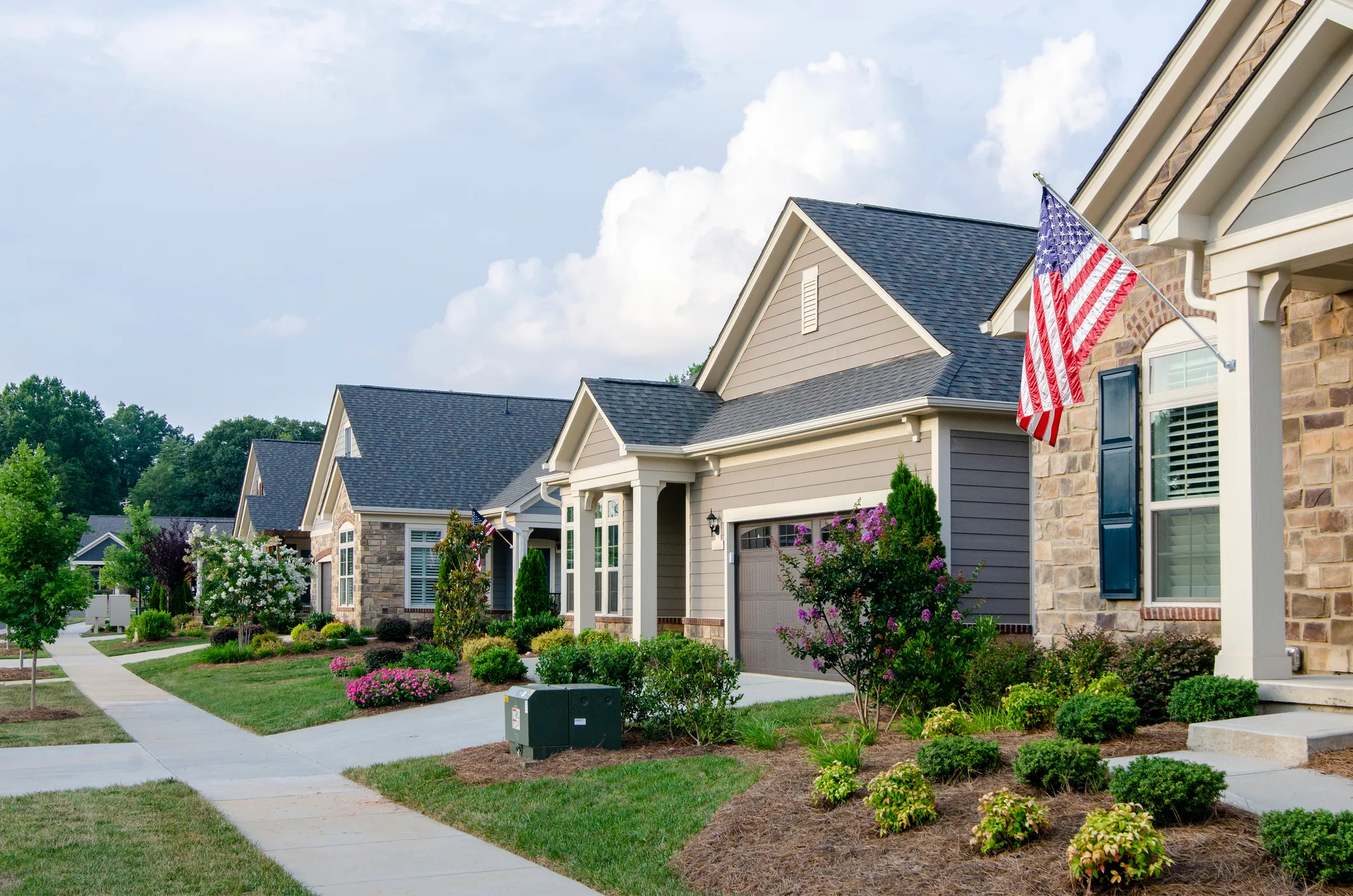 Row of modern suburban houses with manicured lawns and American flags on porches under a partly cloudy sky.