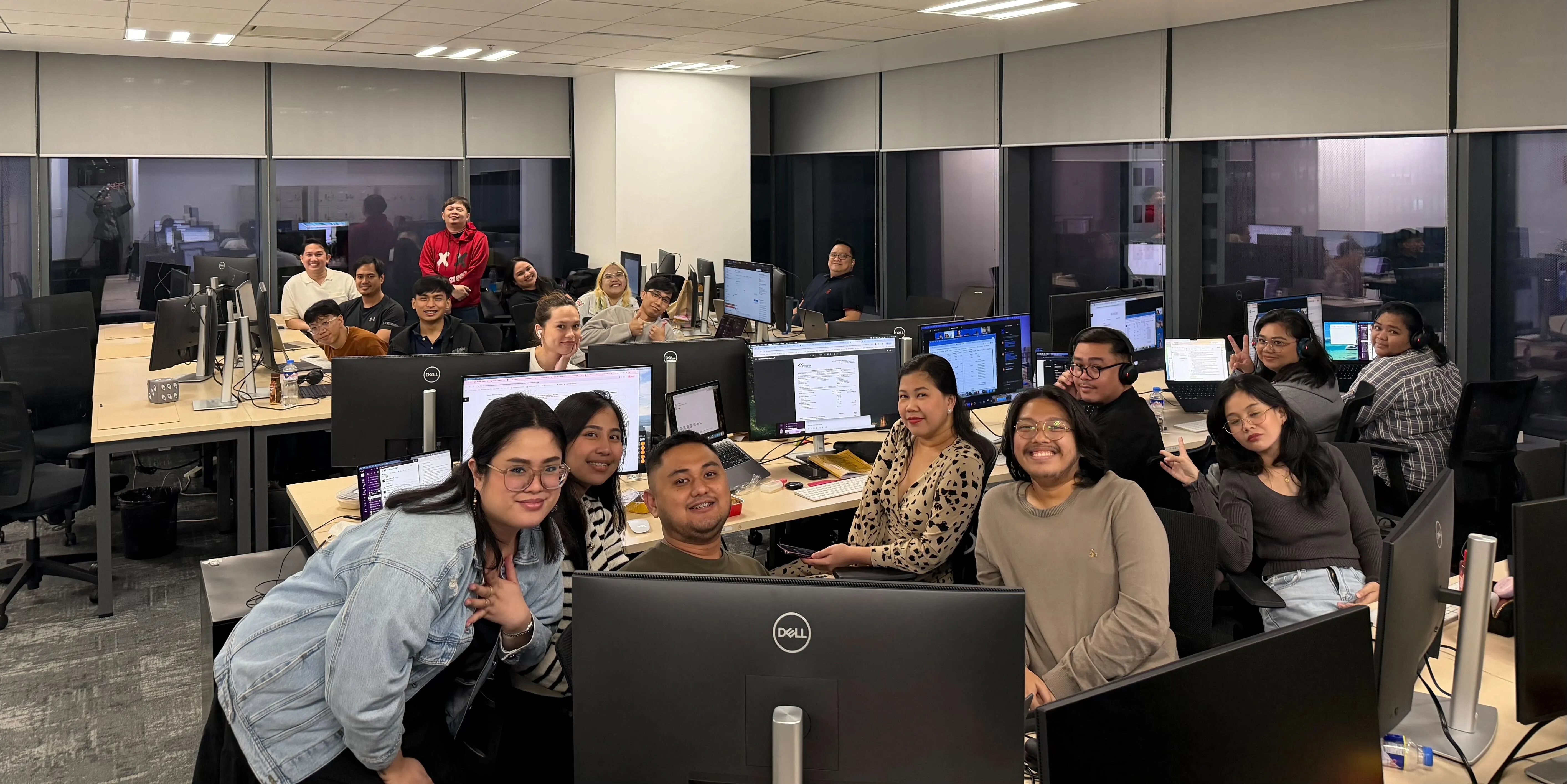 Group of diverse coworkers posing cheerfully in a modern office with computer monitors and desks.