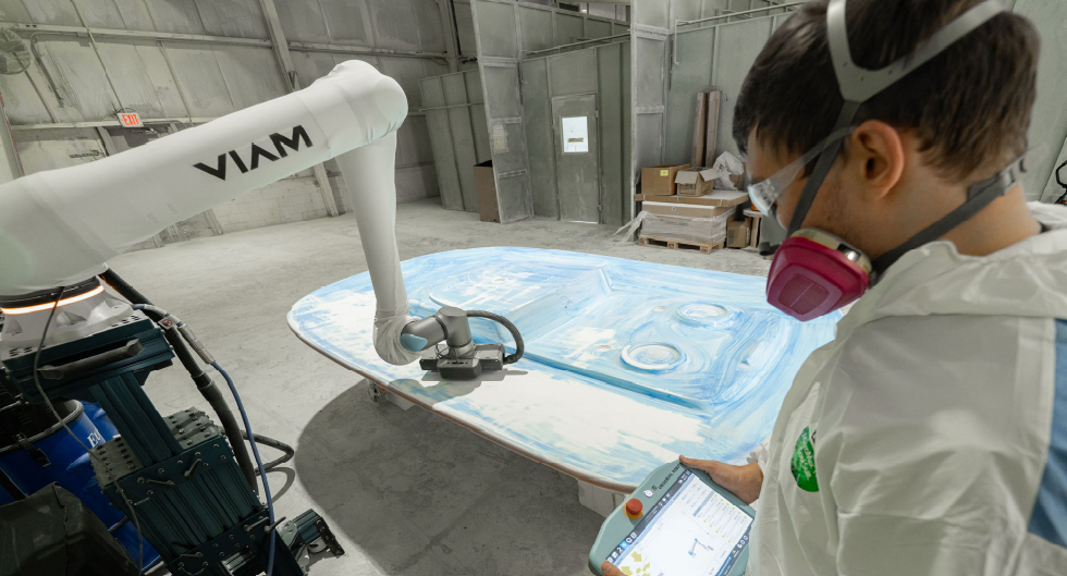 A photo of a robotic arm sanding a boat part while an engineer observes