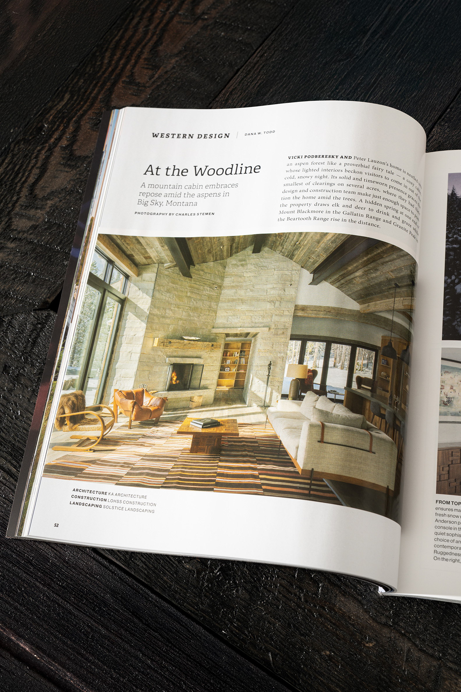 Open magazine on a wooden table featuring an article titled “At the Woodline” with a large photograph of a rustic mountain cabin living room with a stone fireplace and large windows.