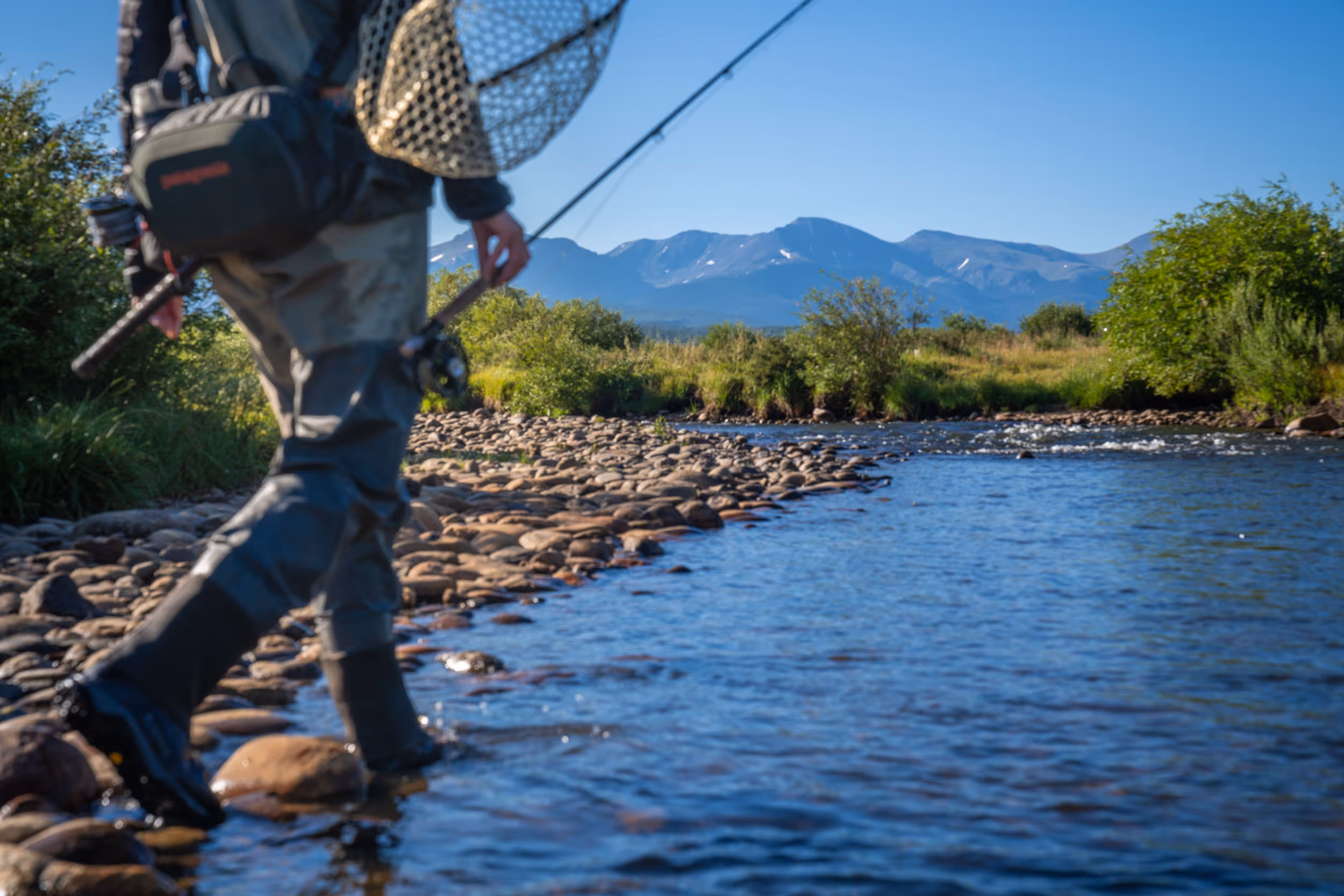 A fly fisherman walking through the clear shallows of a river with their net and fishing rod 