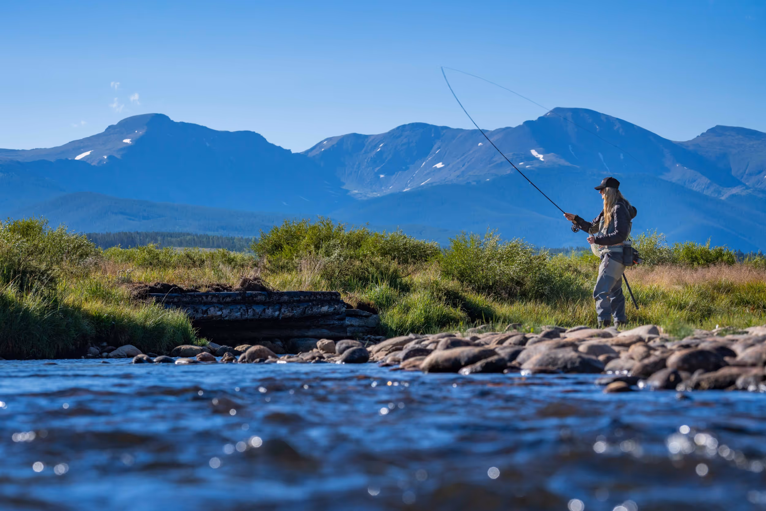 A fly fisher woman casting her line in the sun with a mountain in the background 