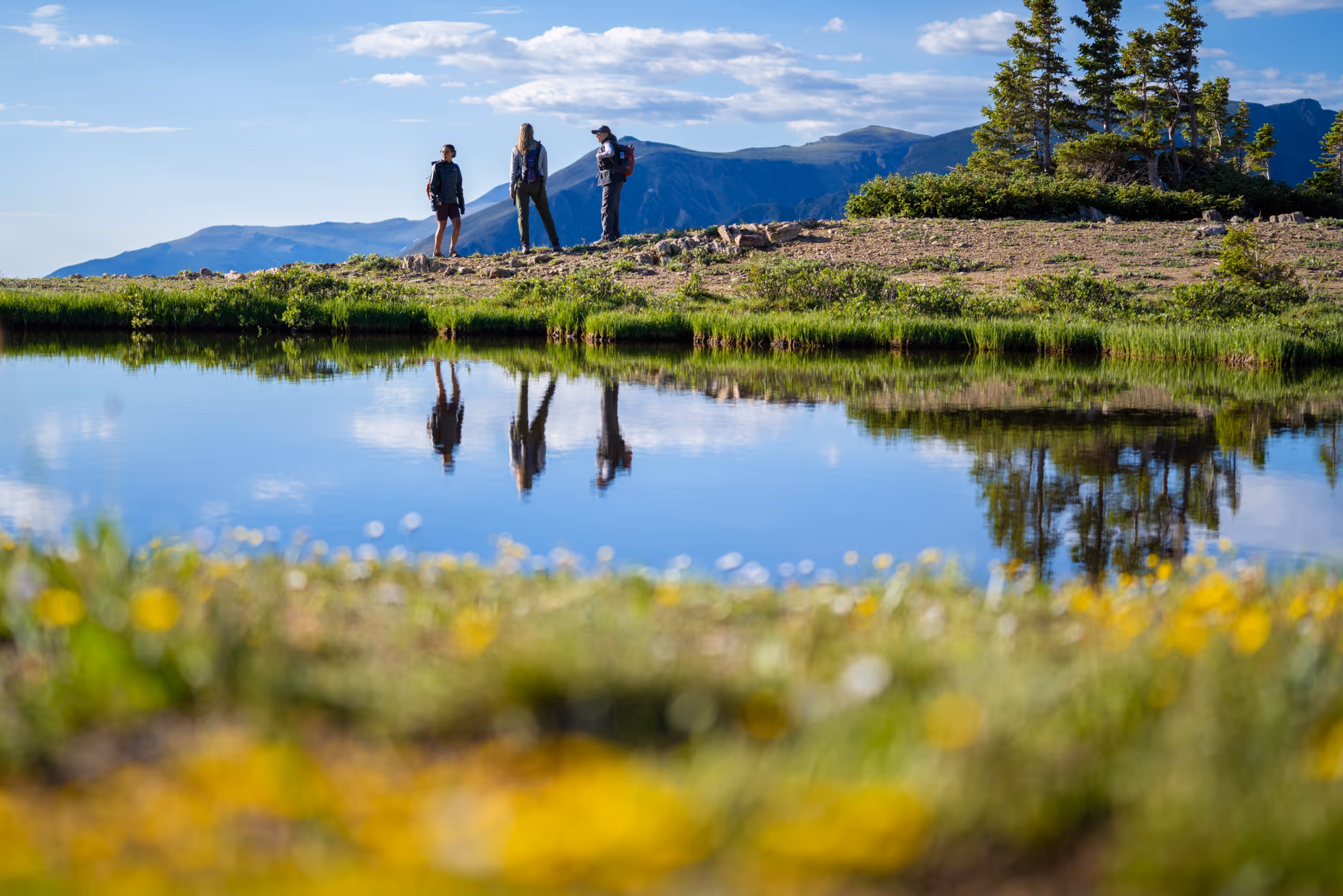 3 hikers standing next to a pond with their reflections in the water
