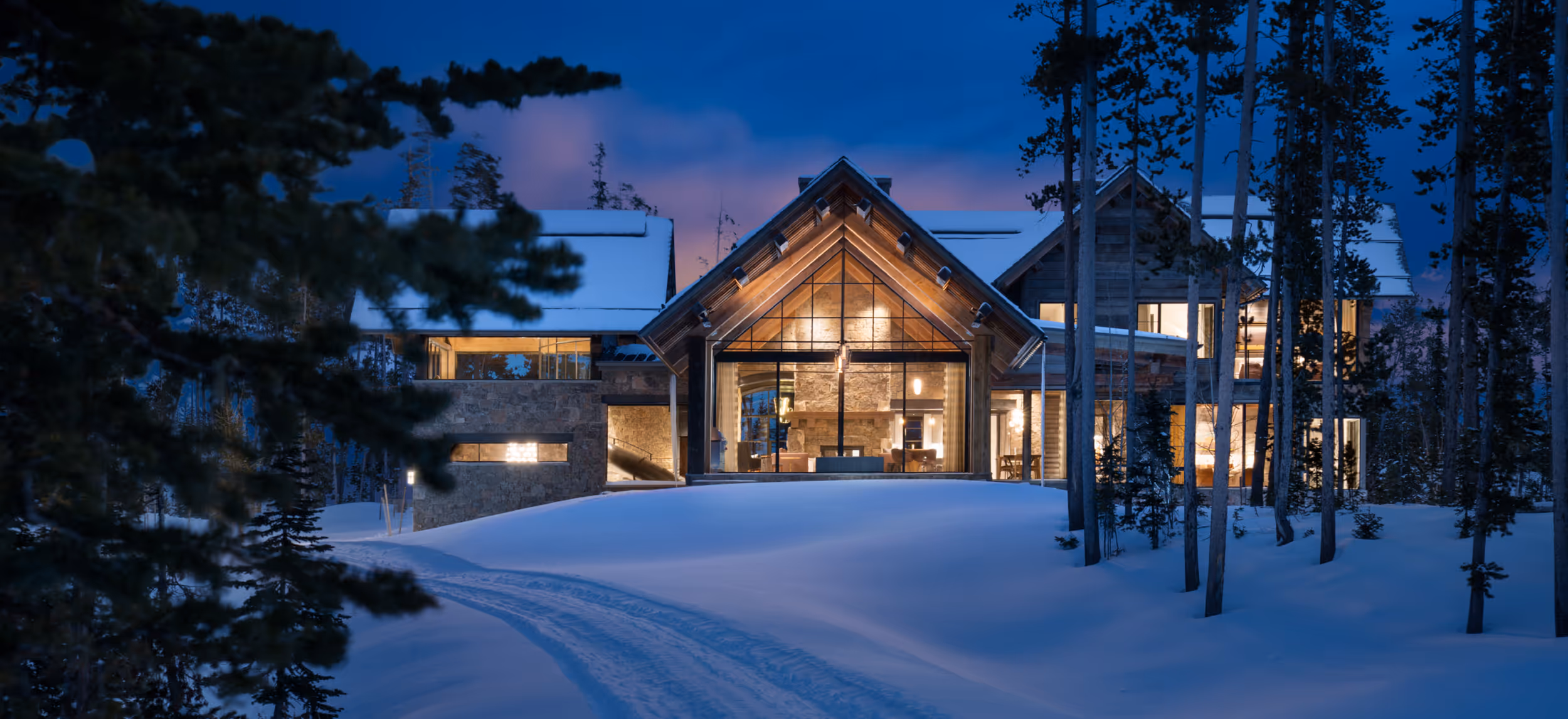 A sunset shot of a  luxurious house with large windows reflecting light on the snow and forest in the foreground. 