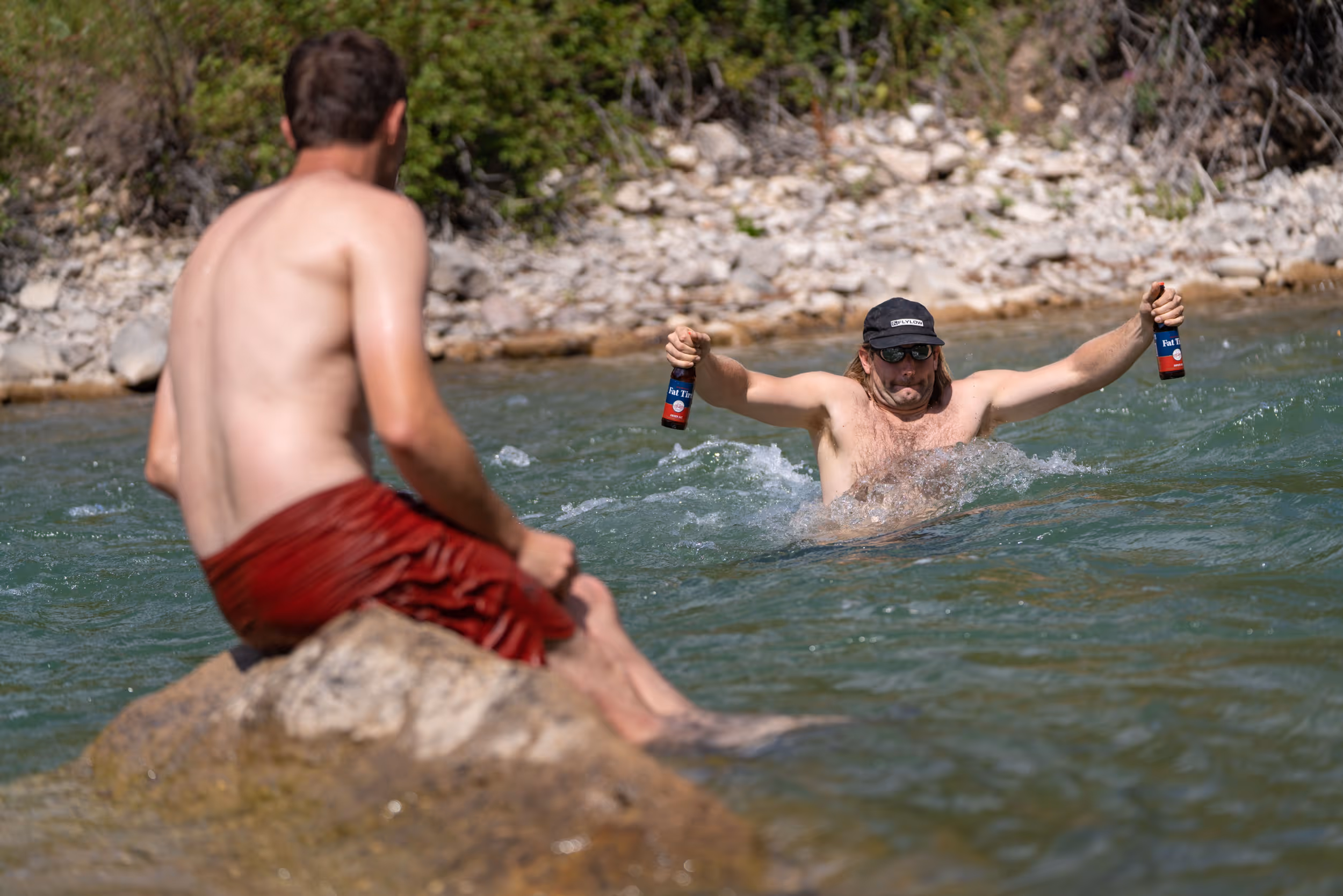 A man making a brave river crossing while keeping 2 beers out of the water so he can bring a beer to his buddy chilling on a rock
