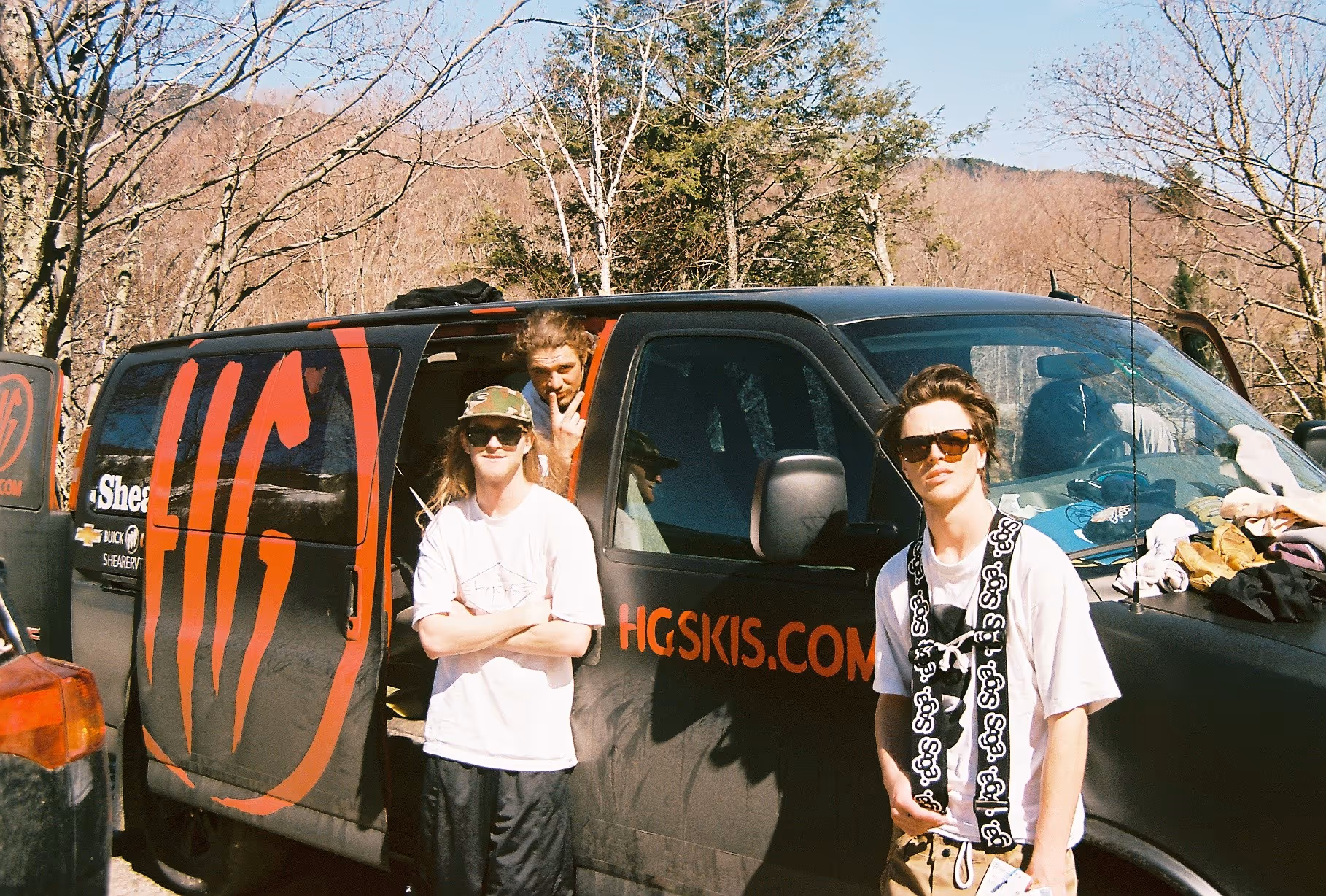 A few skiers hanging in front of a black van with orange HG ski decal on the exterior 