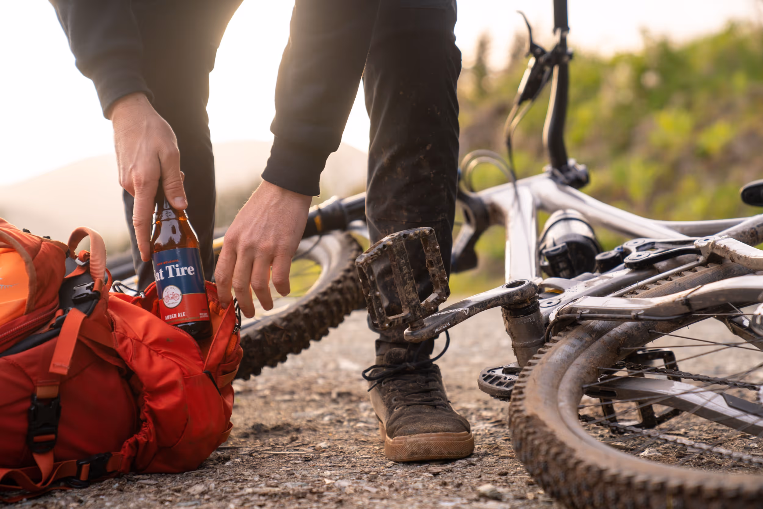 A muddy mountain biker with their bike laying on the ground reaching into their pack for a snack 