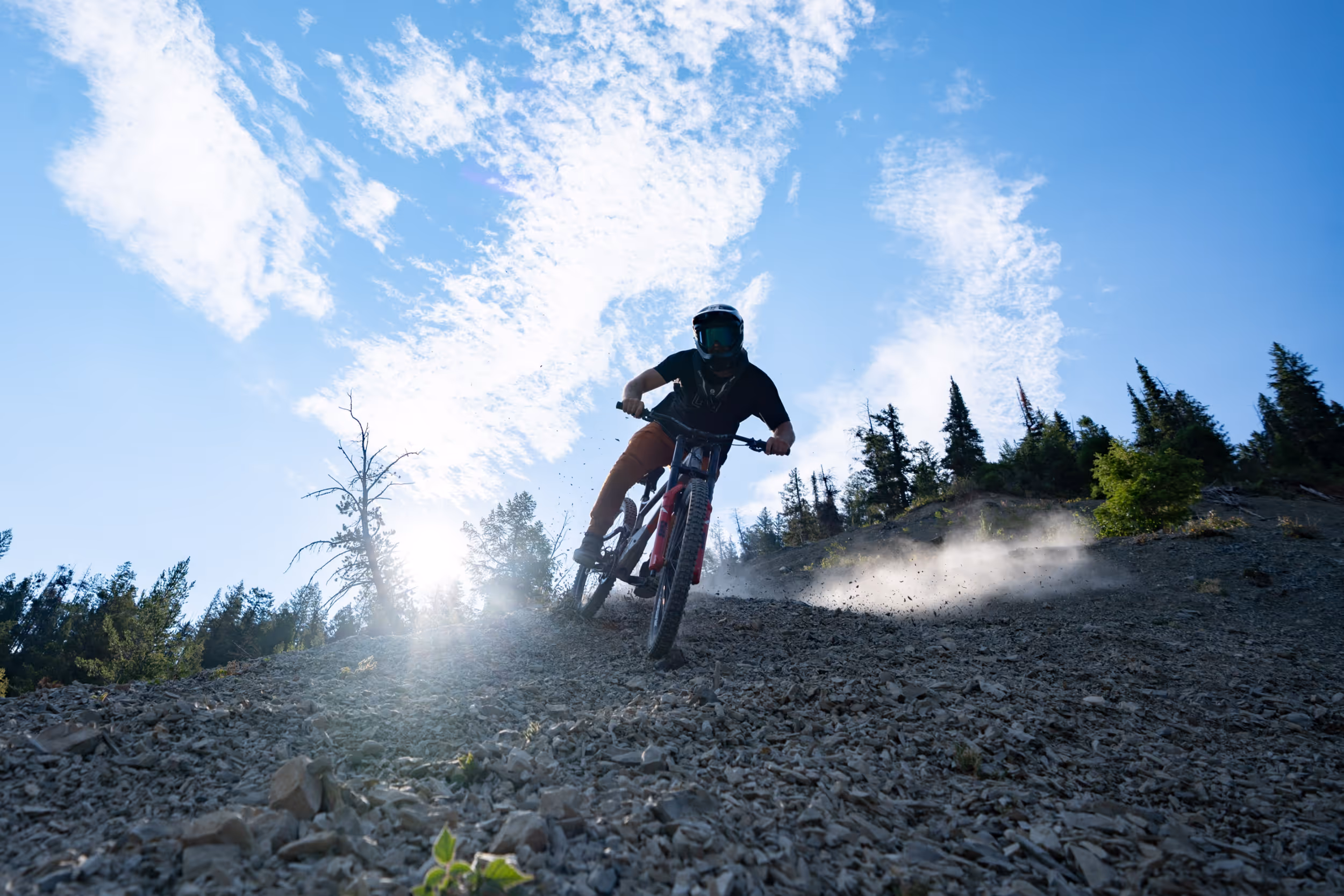 Looking uphill at a mountain biker riding down towards the camera with a bright sky behind him 
