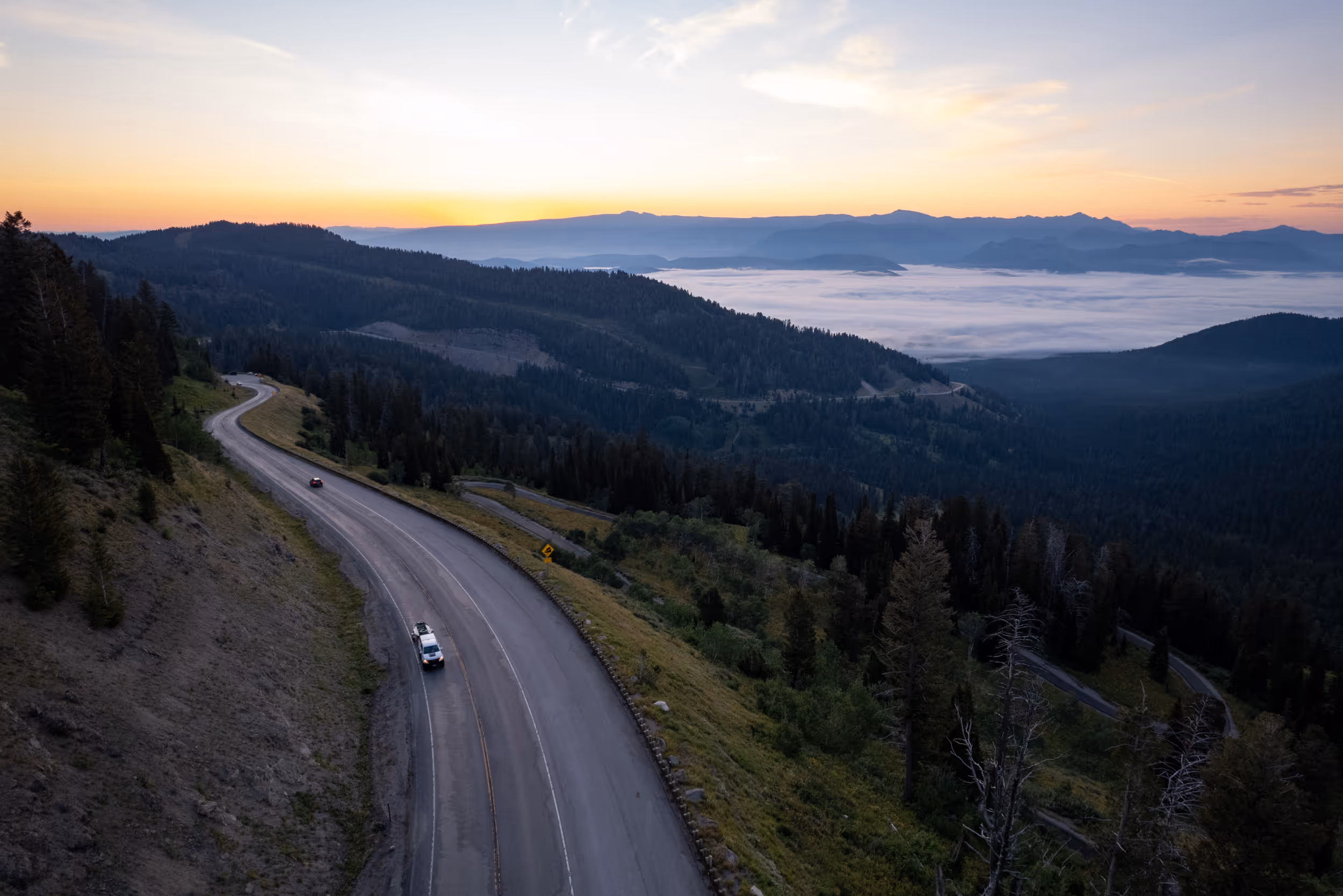 A sunset landscape view of a wooded highway with some cloud cover lower in the valley 