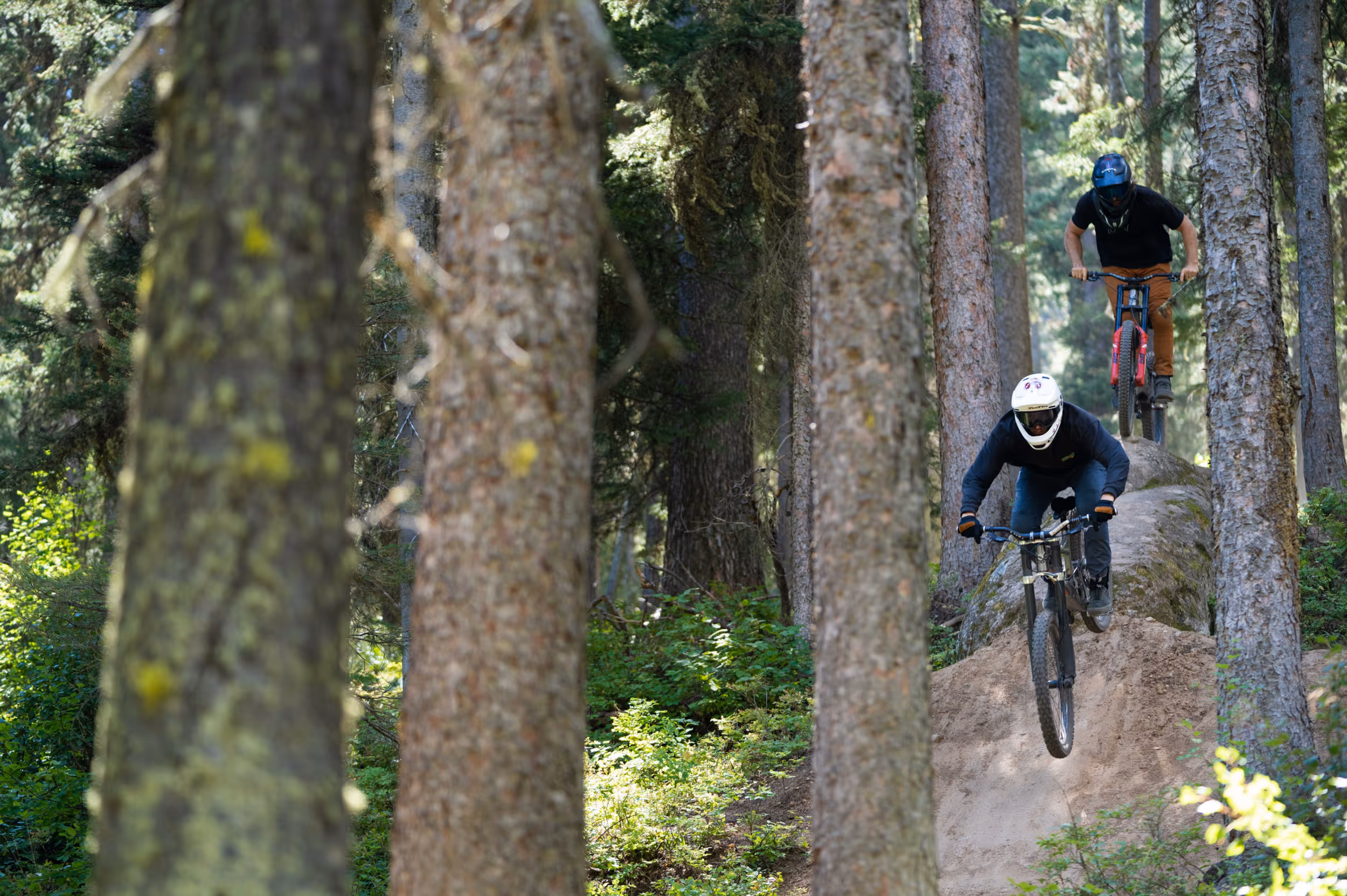 2 mountain bikers riding through a tricky rock feature on a forested trail 