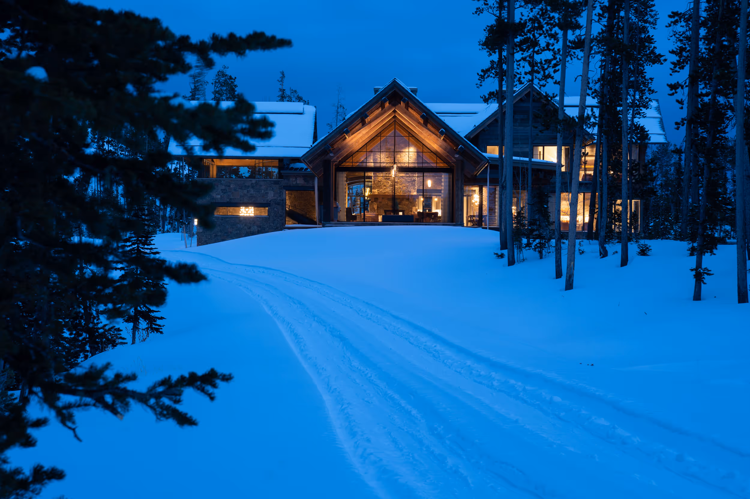 A sunset shot of a  luxurious house with large windows reflecting light on the snow and forest in the foreground. 