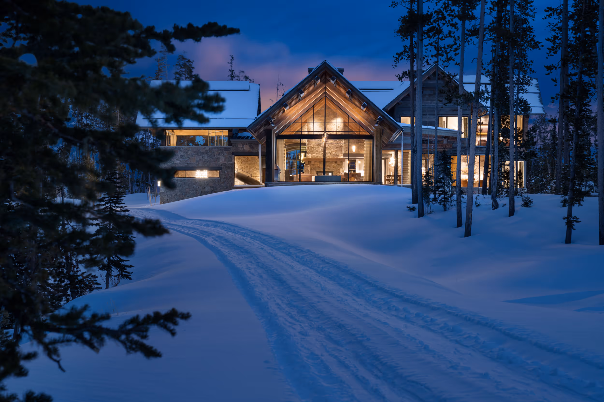 A sunset shot of a  luxurious house with large windows reflecting light on the snow and forest in the foreground. 