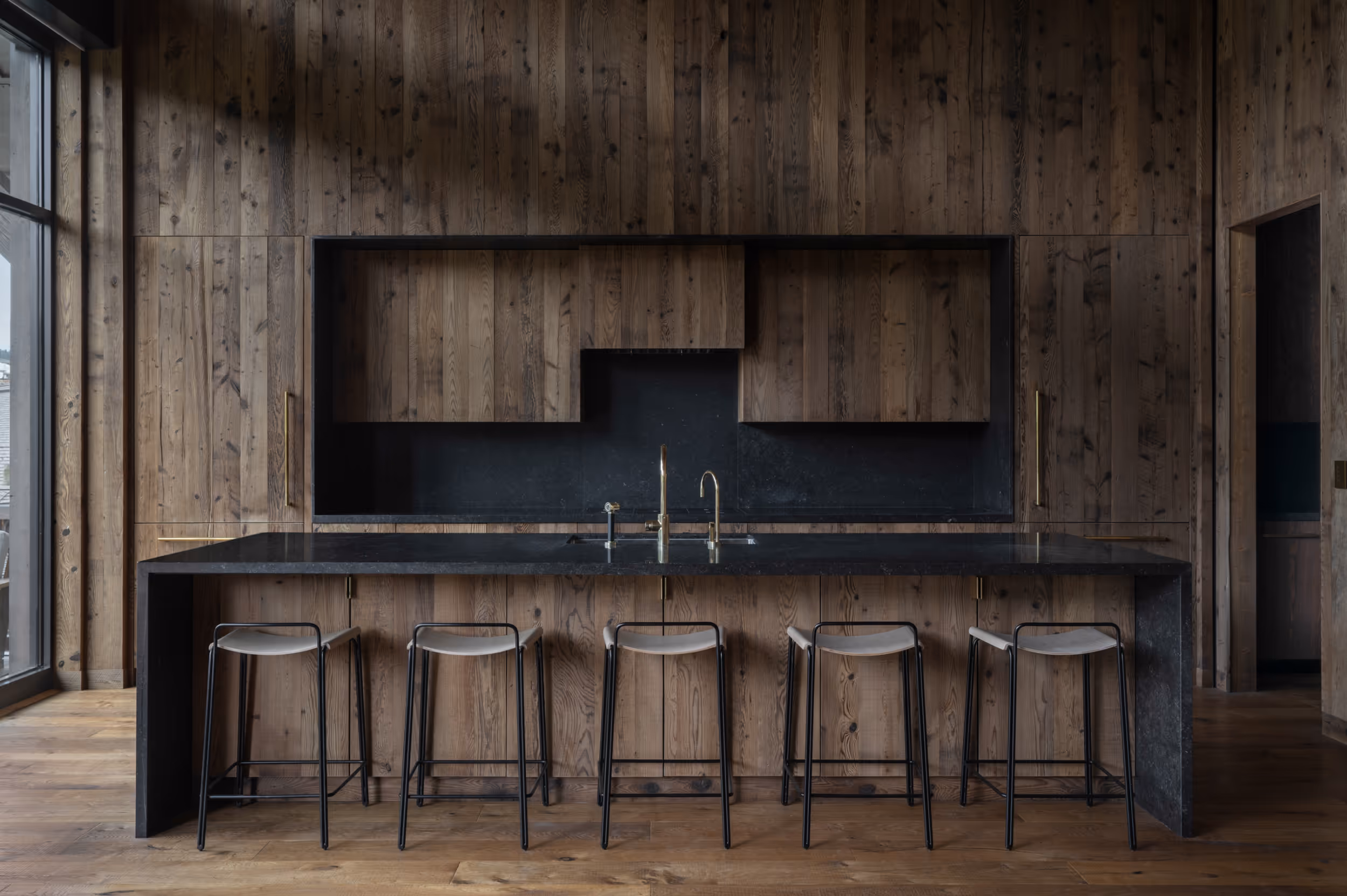 A modern kitchen with large windows letting in a lot of light that juxtaposes the darker wood interior and black marble counter tops  