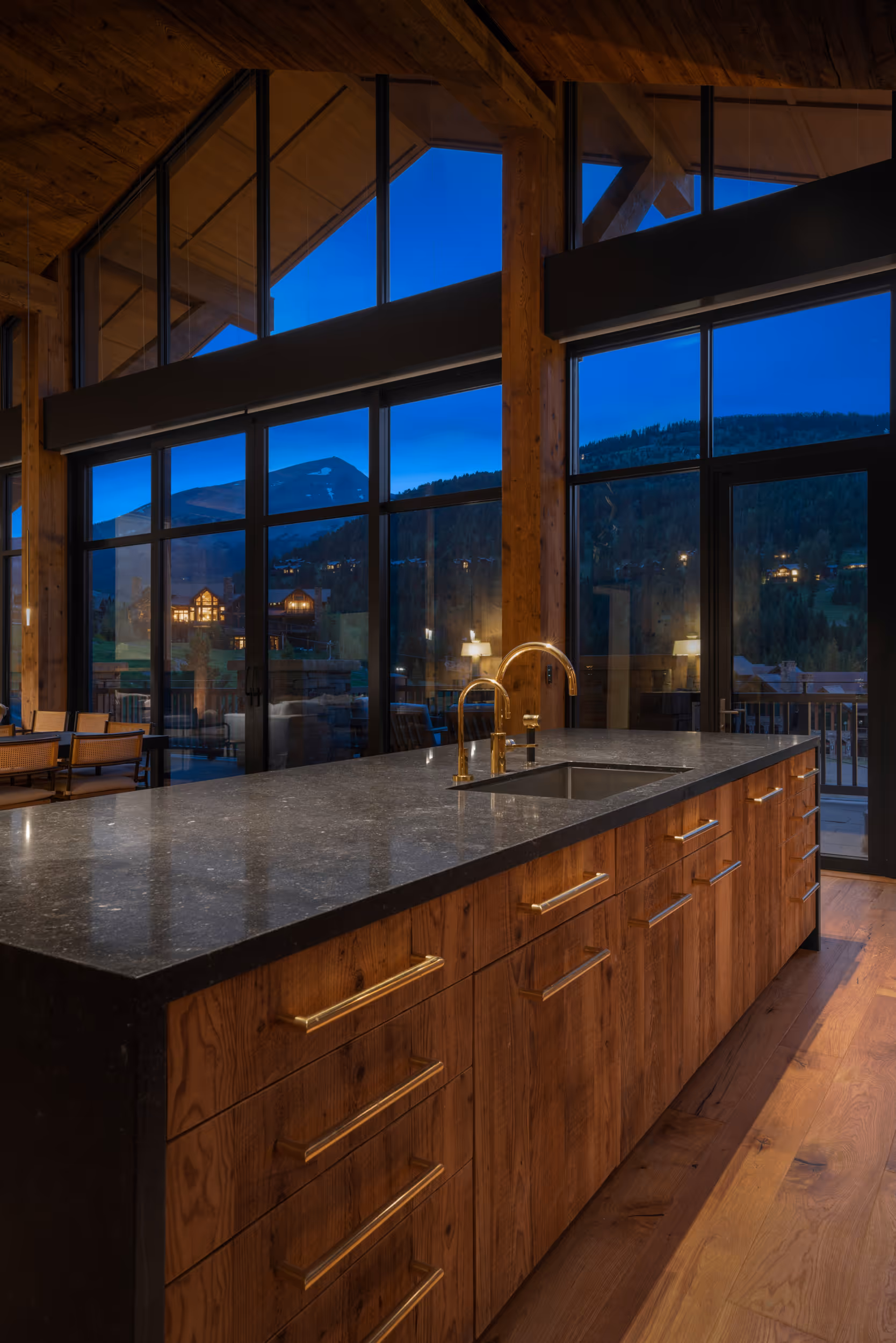 A luxurious modern kitchen with large windows and a dark wooden interior, and lots of warm lighting  