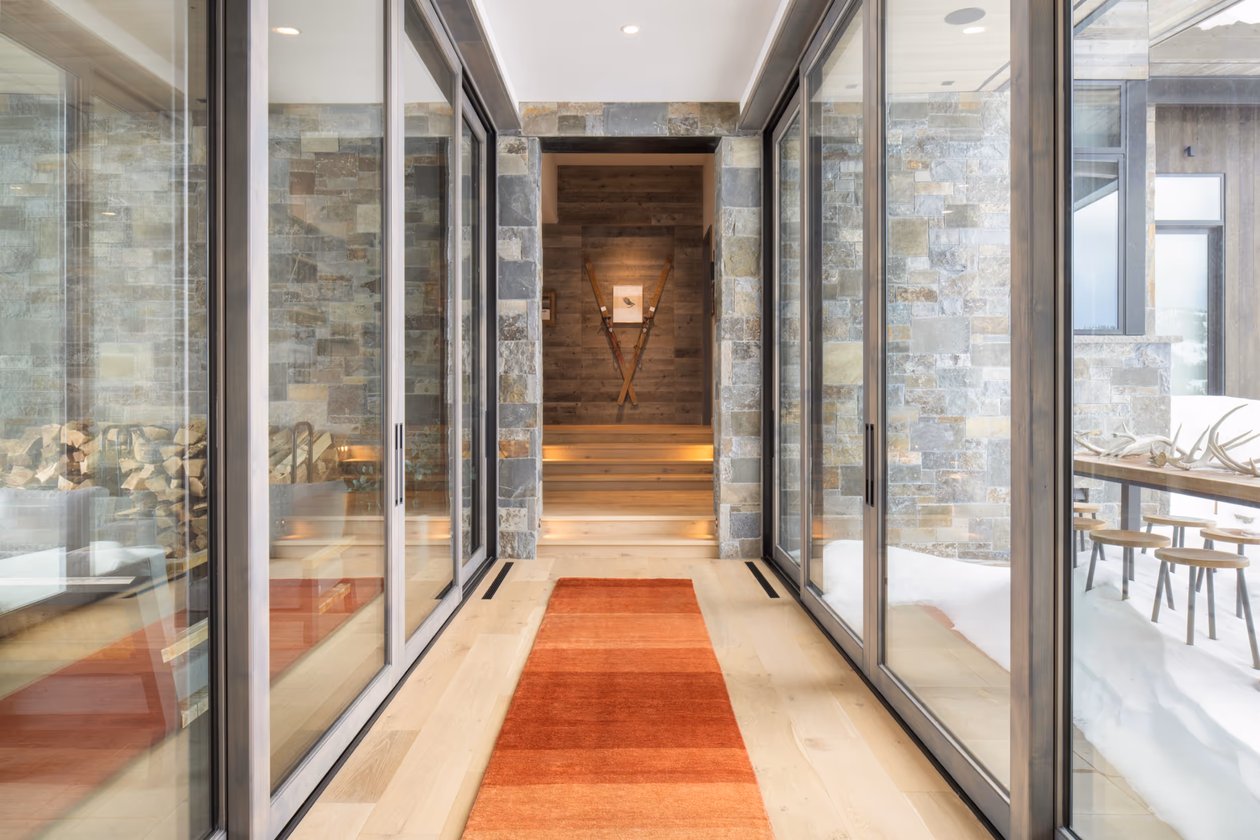 An elegant, modern hall way surrounded by glass walls, with an orange rug, and skis mounted on the far wall 