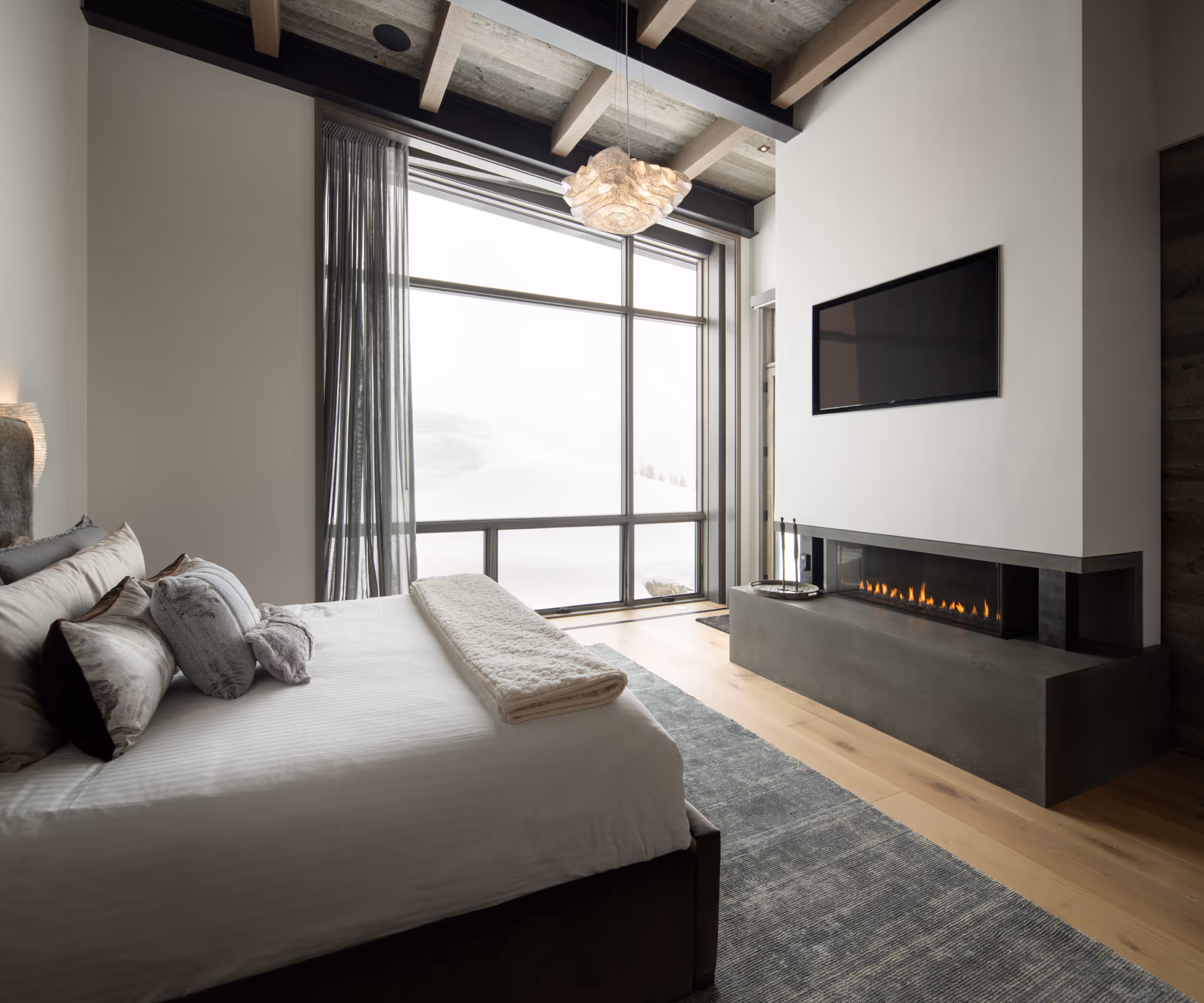 A large bedroom with a fireplace, luxurious bed, and a wall that's made almost entirely from windows 