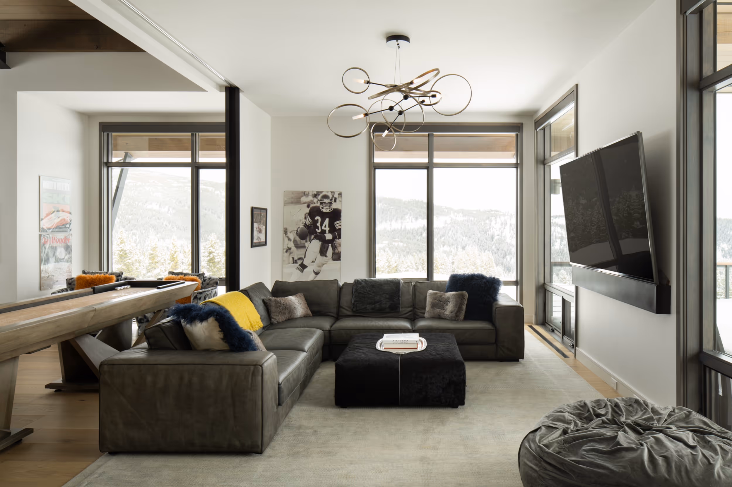 A large tv room connected to a game room with a large sectional couch, tv, and a lovely geometric light fixture 