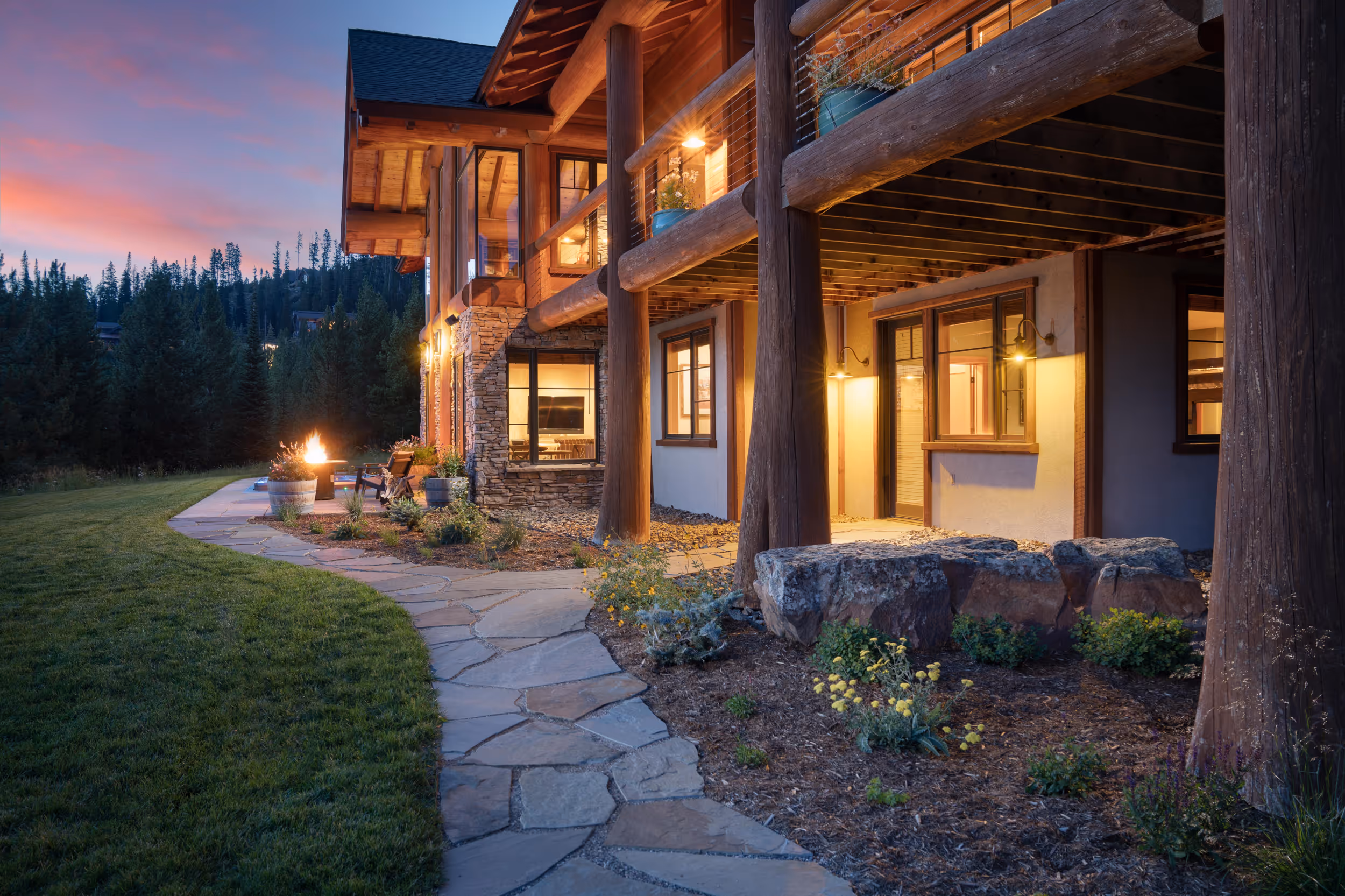 A sunset photo of the lower section of a luxurious stone and log cabin showing the well lit interior and exterior of the house